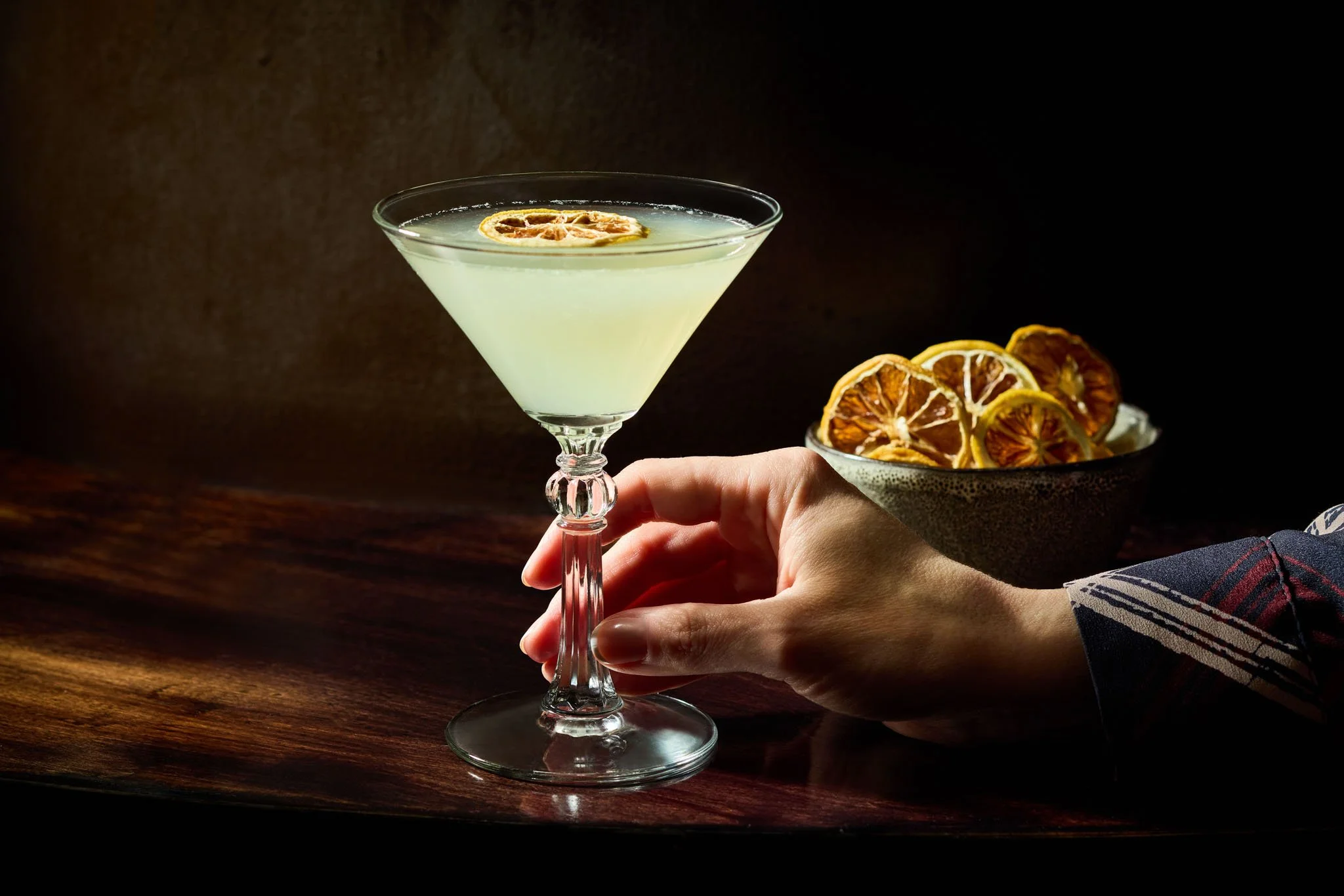 A hand holding a cocktail glass with a light-colored drink and a lemon slice garnish, with a bowl of dried lemon slices in the background on a dark wooden surface.