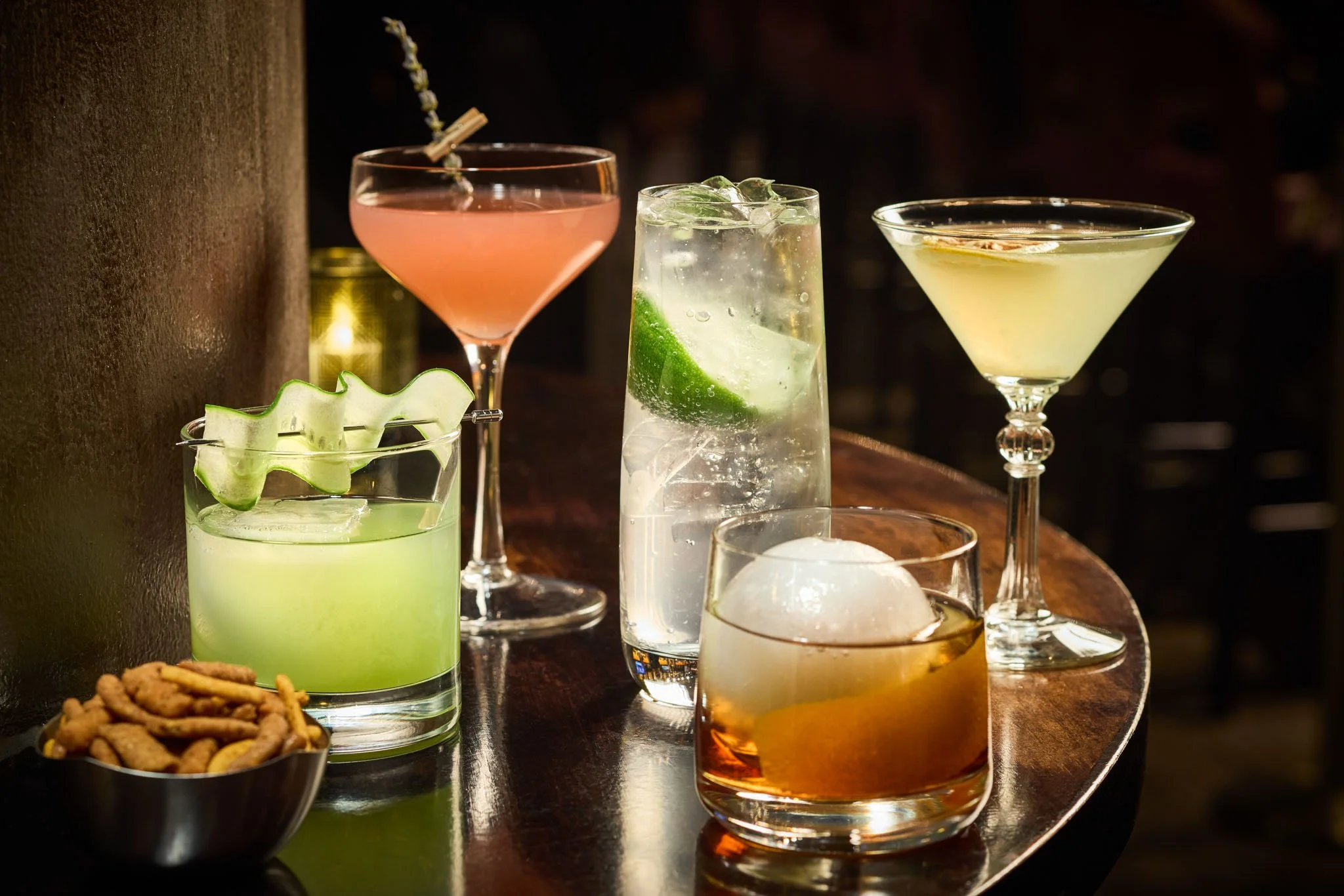 Six different cocktails with various colors and glass styles on a dark wooden bar table, with a bowl of mixed nuts in the foreground.