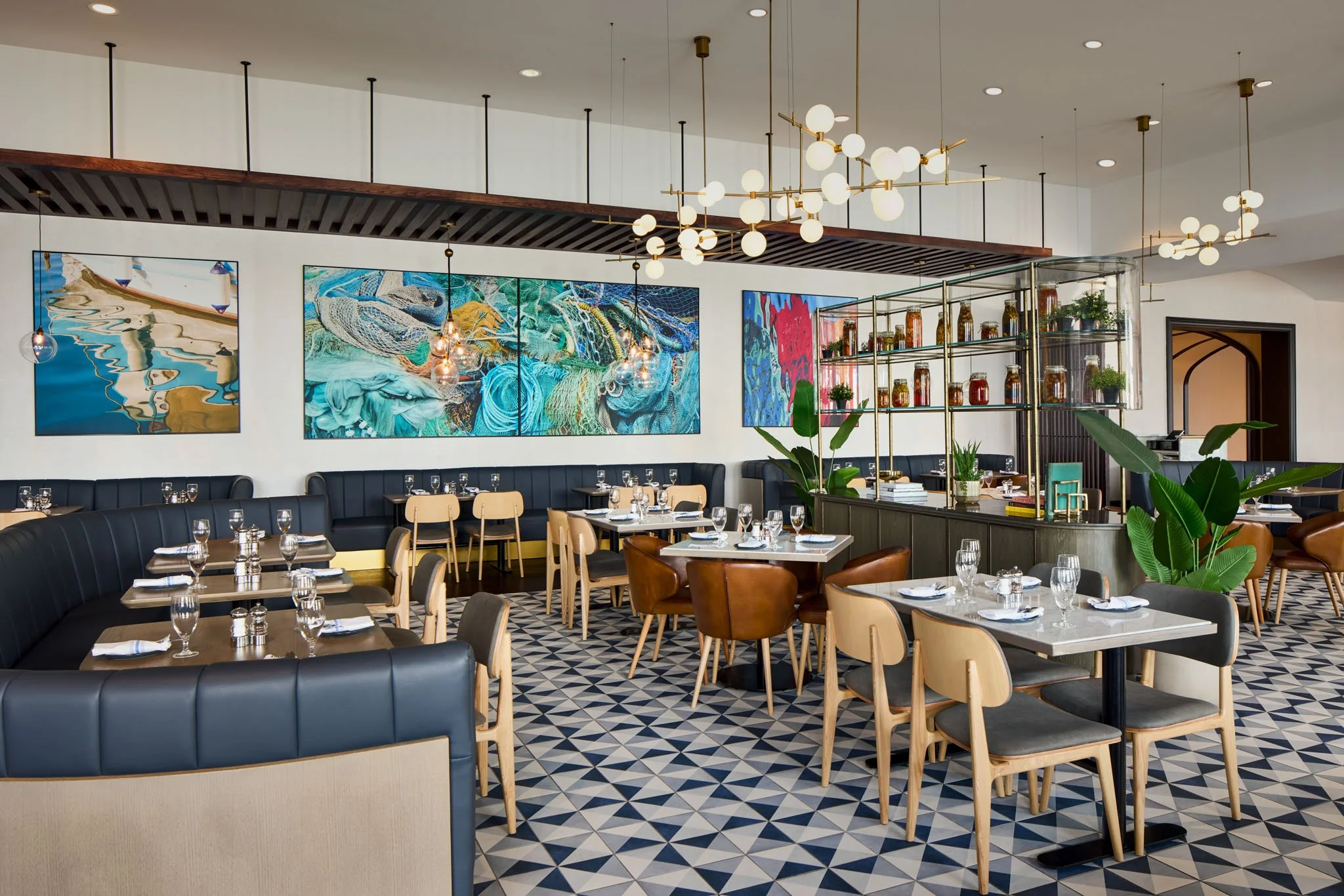 Modern restaurant interior with black leather booth seating, wooden chairs, patterned tile floor, colorful abstract artwork on white walls, and decorative lighting fixtures.