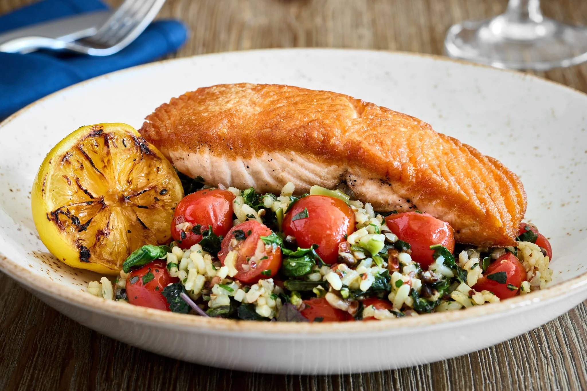 Cooked salmon fillet with grilled lemon and a side of tomato and greens rice salad on a white plate.