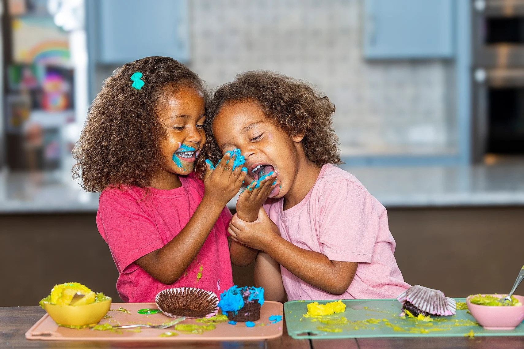 Two young girls sharing a cake at a birthday party, covered in blue and yellow frosting, with cake and bowls on the table.