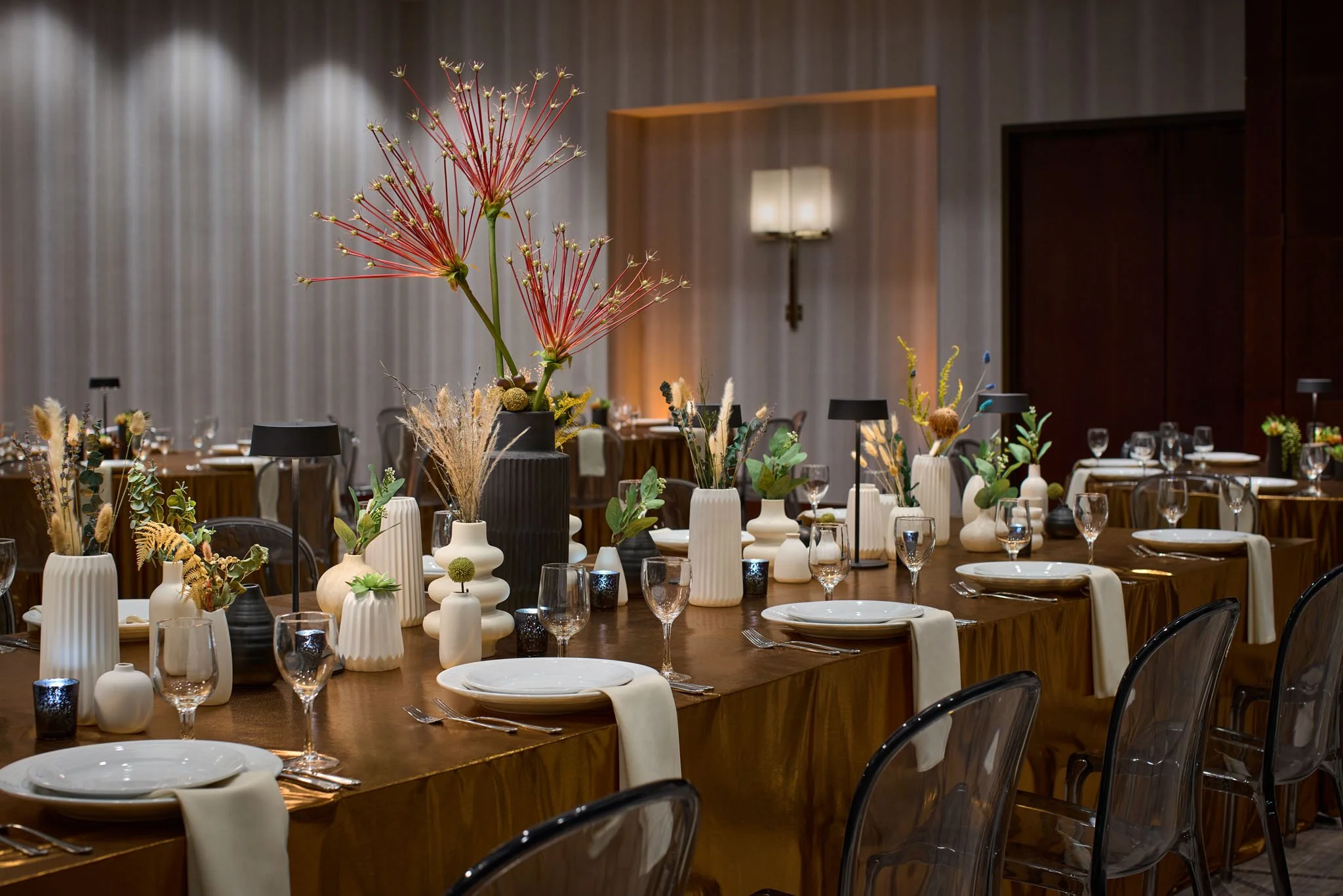 Elegant dining table set with white plates, glasses, napkins, and floral centerpieces in vases, in a decorated event space.