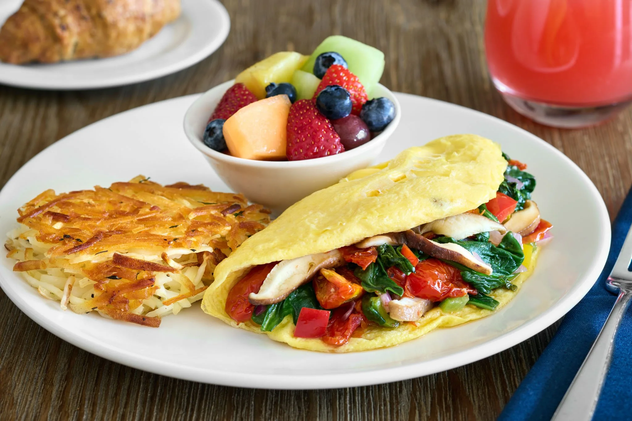 Omelette with vegetables, hash browns, fresh fruit salad, and a glass of pink beverage on a white plate and wooden table.