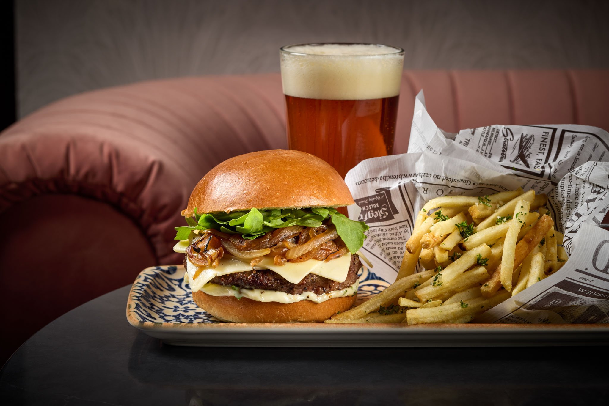A plate with a cheeseburger with lettuce, onions, cheese, and a beef patty, alongside a side of French fries garnished with parsley, a pint of amber beer, and a wrapped serving of fries on a napkin. The Noble Fox in Le Meridien Arlington, VA