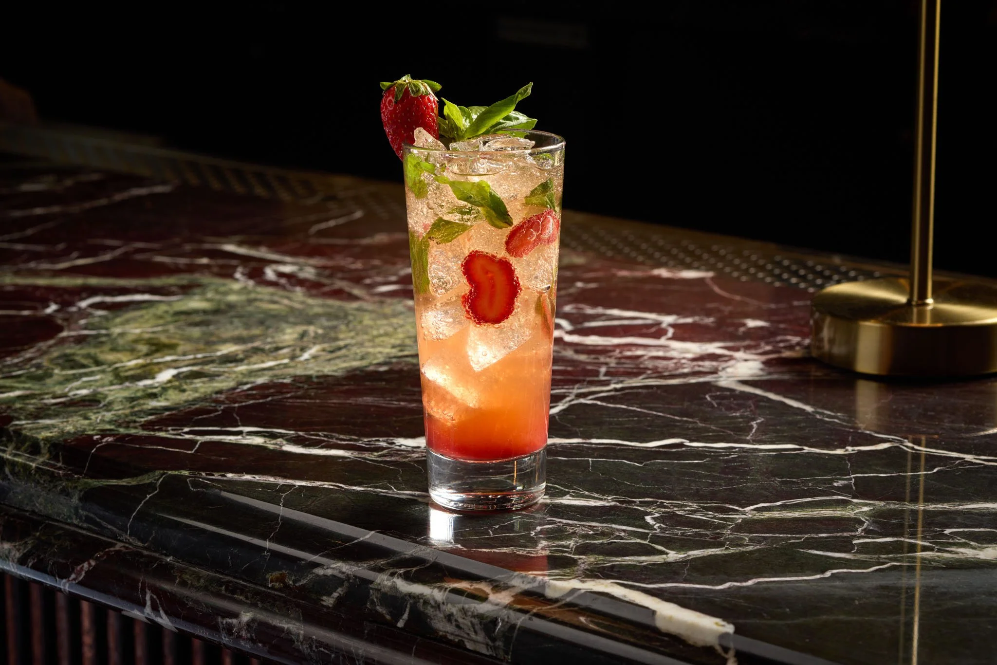A tall glass of strawberry and basil-infused drink with ice, garnished with a strawberry, basil leaves, and a straw, on a marble table with a dark background.