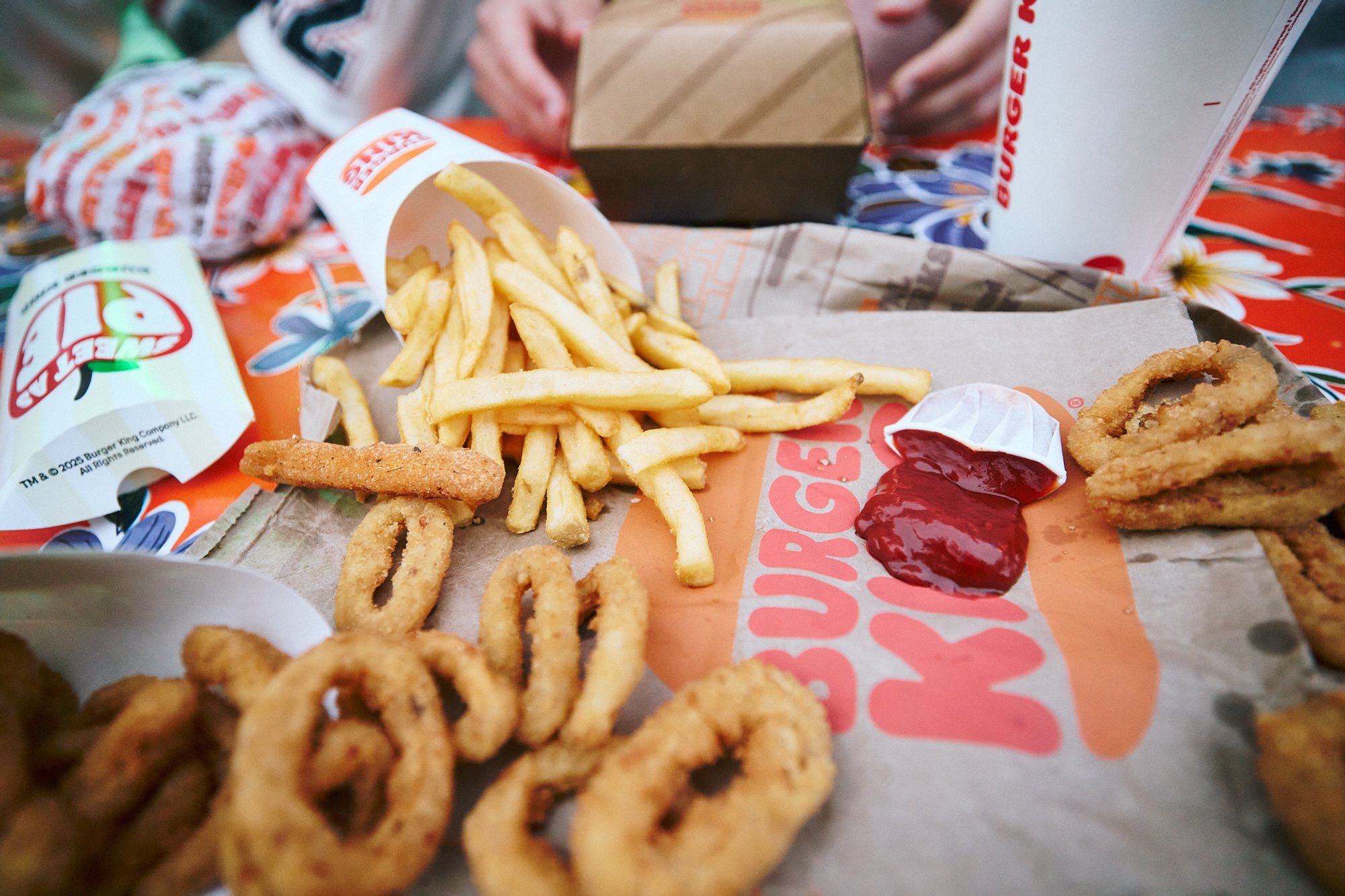 Burger King food tray with curly fries, French fries, onion rings, ketchup, a fried chicken strip, and a burger in the background. Burger box and soda cup with visible Burger King branding.
