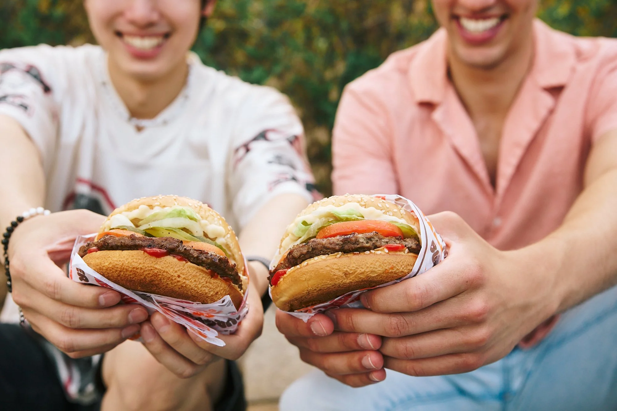 Two people holding Burger King Whoppers in their hands outdoors during daytime, smiling, with blurred greenery in the background.
