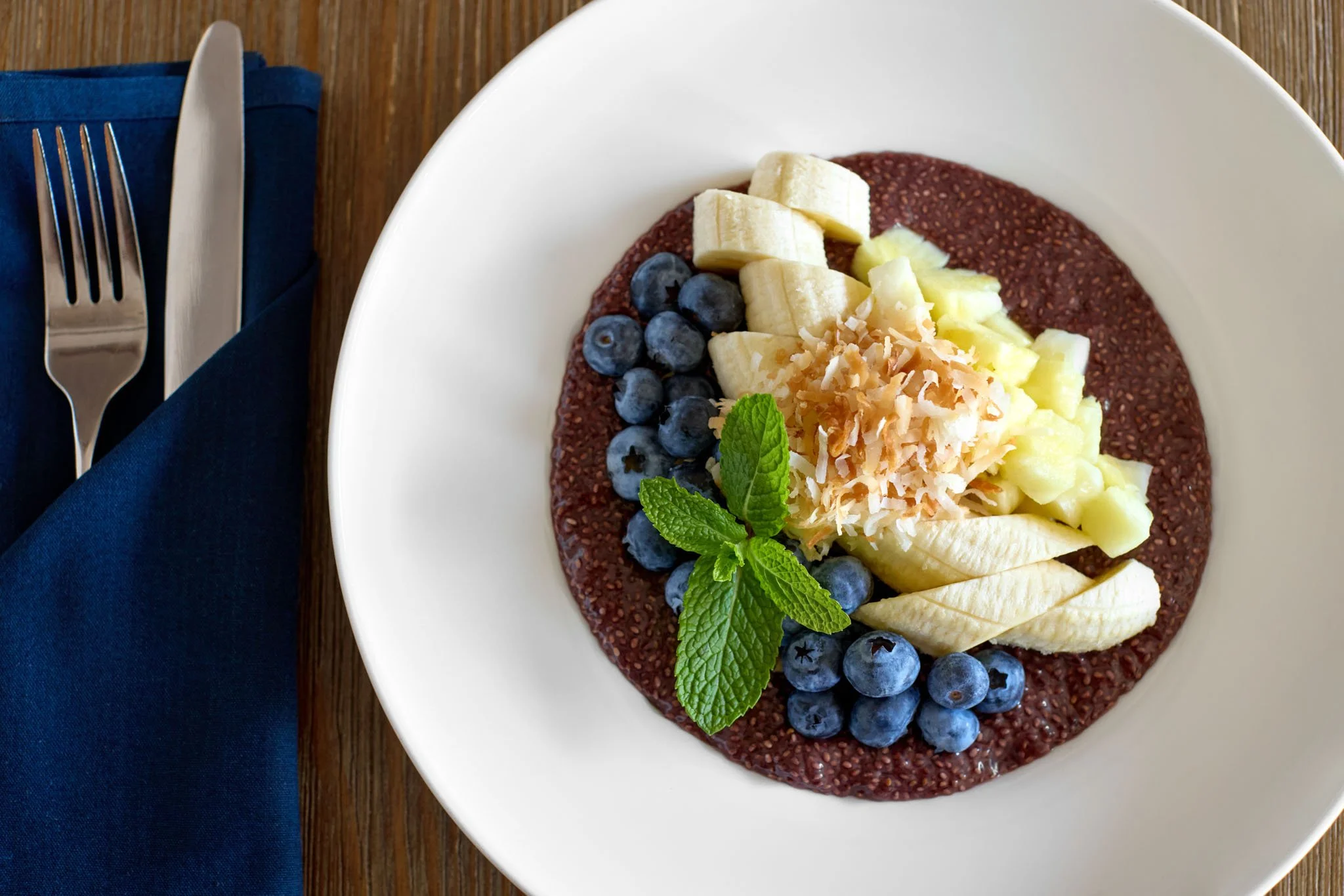 A white bowl filled with a berry chia pudding topped with sliced banana, blueberries, diced honeydew melon, shredded coconut, and fresh mint leaves, placed on a wooden table with a blue napkin and a fork and knife.