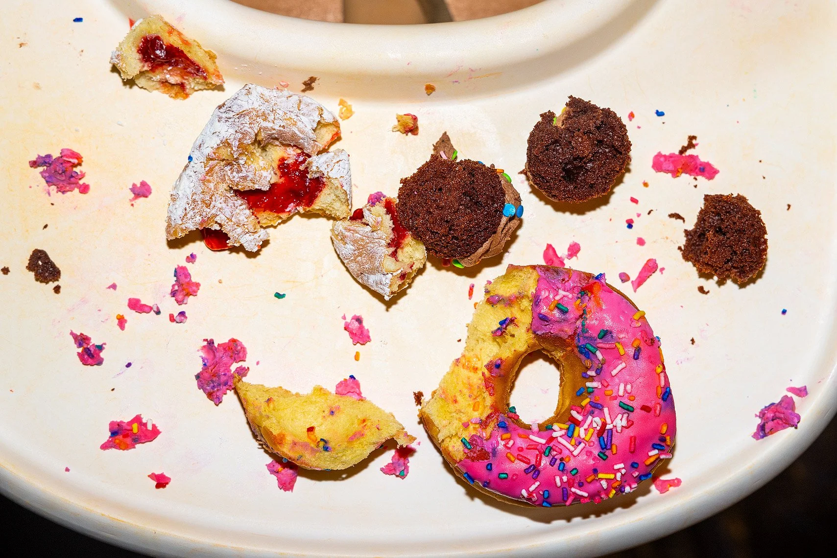 Broken donut with pink frosting and sprinkles along with cookie crumbs, chocolate truffles, and other snack remnants on a white plate.