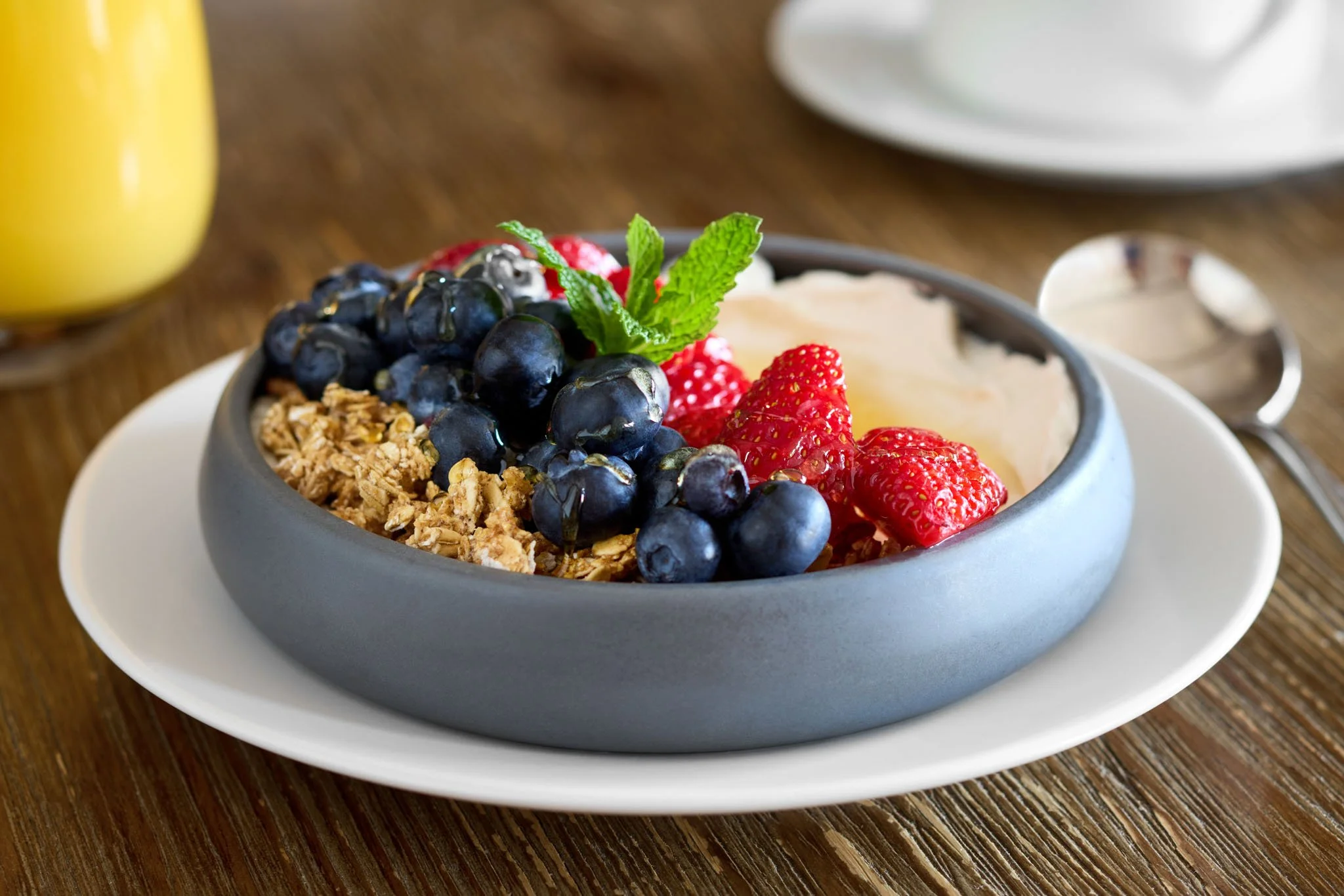 A bowl of yogurt topped with blueberries, strawberries, granola, a mint leaf, and a few red raspberries, on a white plate on a wooden table.