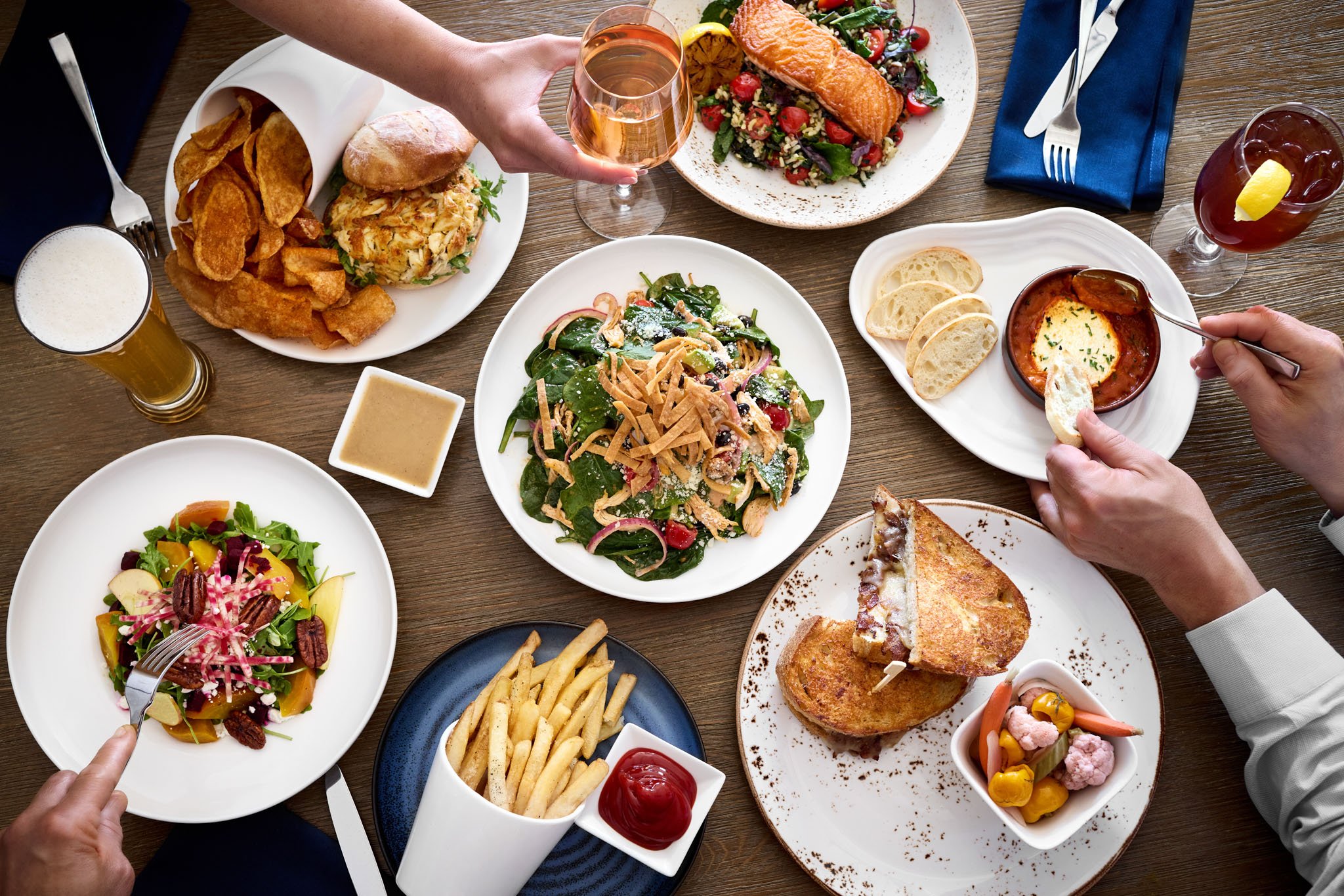 Various plates of food including salads, soup, and fried dishes on a wooden table, with hands holding drinks and utensils.