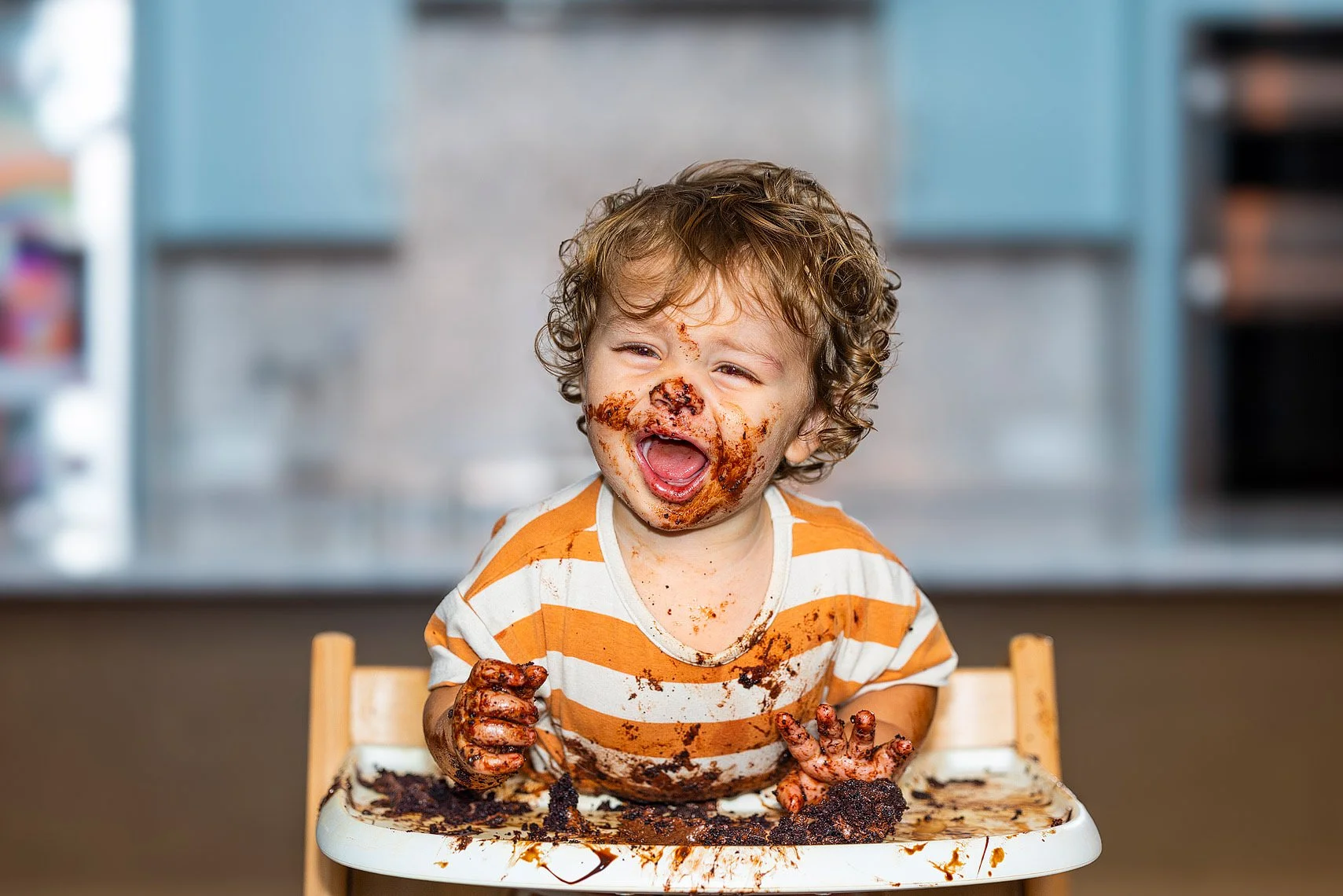 Child with curly hair and a striped orange and white shirt sitting in a high chair, covered in chocolate and smiling