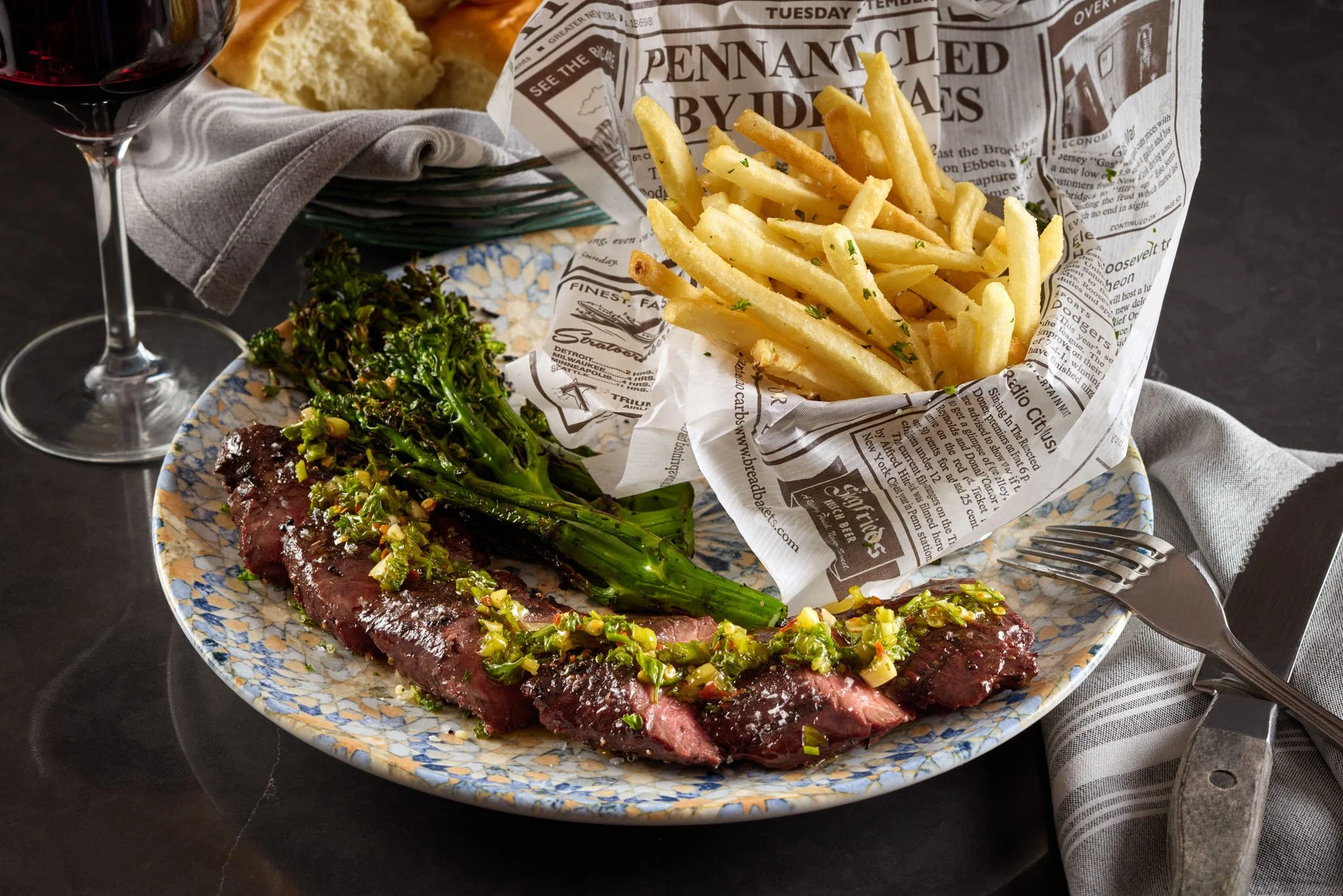 A plate of food featuring a grilled steak topped with chopped green onions, accompanied by a cooked leafy green vegetable, with a side of French fries, and a glass of red wine. Noble Fox, Le Meridien Arlington, VA