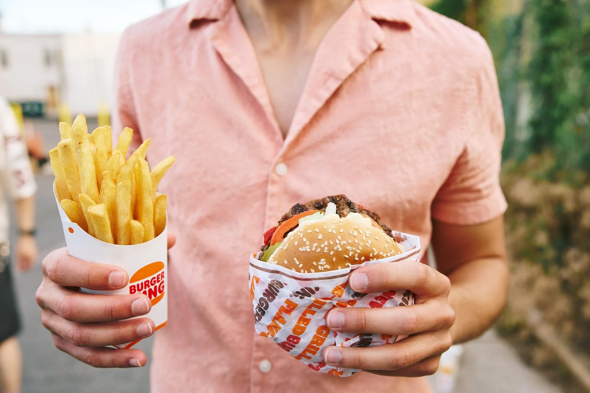 Person holding a container of Burger King French fries in one hand and a Whopper in the other, in an outdoor setting.