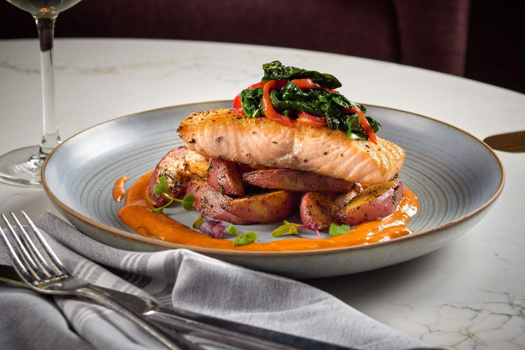 A plated dish featuring a piece of cooked salmon topped with sautéed greens and red peppers, served over roasted potatoes and surrounded by an orange sauce. A glass of white wine. Noble Fox Le Meridien Arlington VA