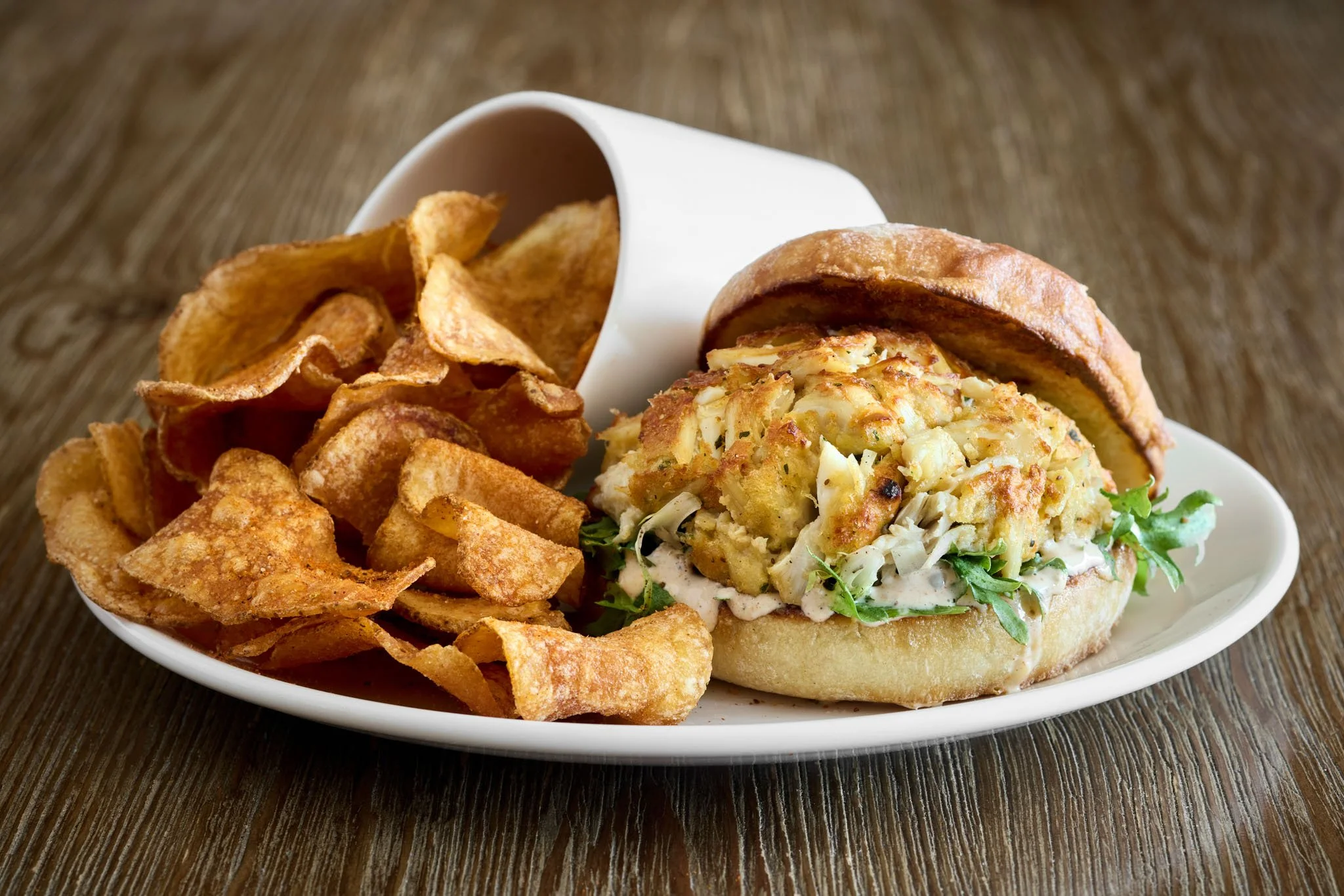 A plate with potato chips and a fish sandwich in a bun on a wooden surface.
