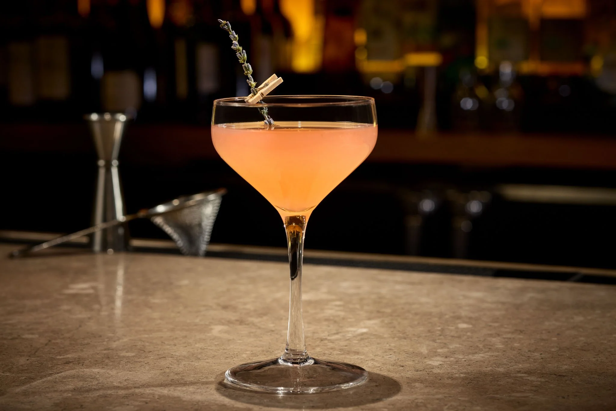 A pink cocktail with a sprig of lavender garnished with a small wooden clothespin in a stemmed glass on a marble countertop, with bar tools in the background.