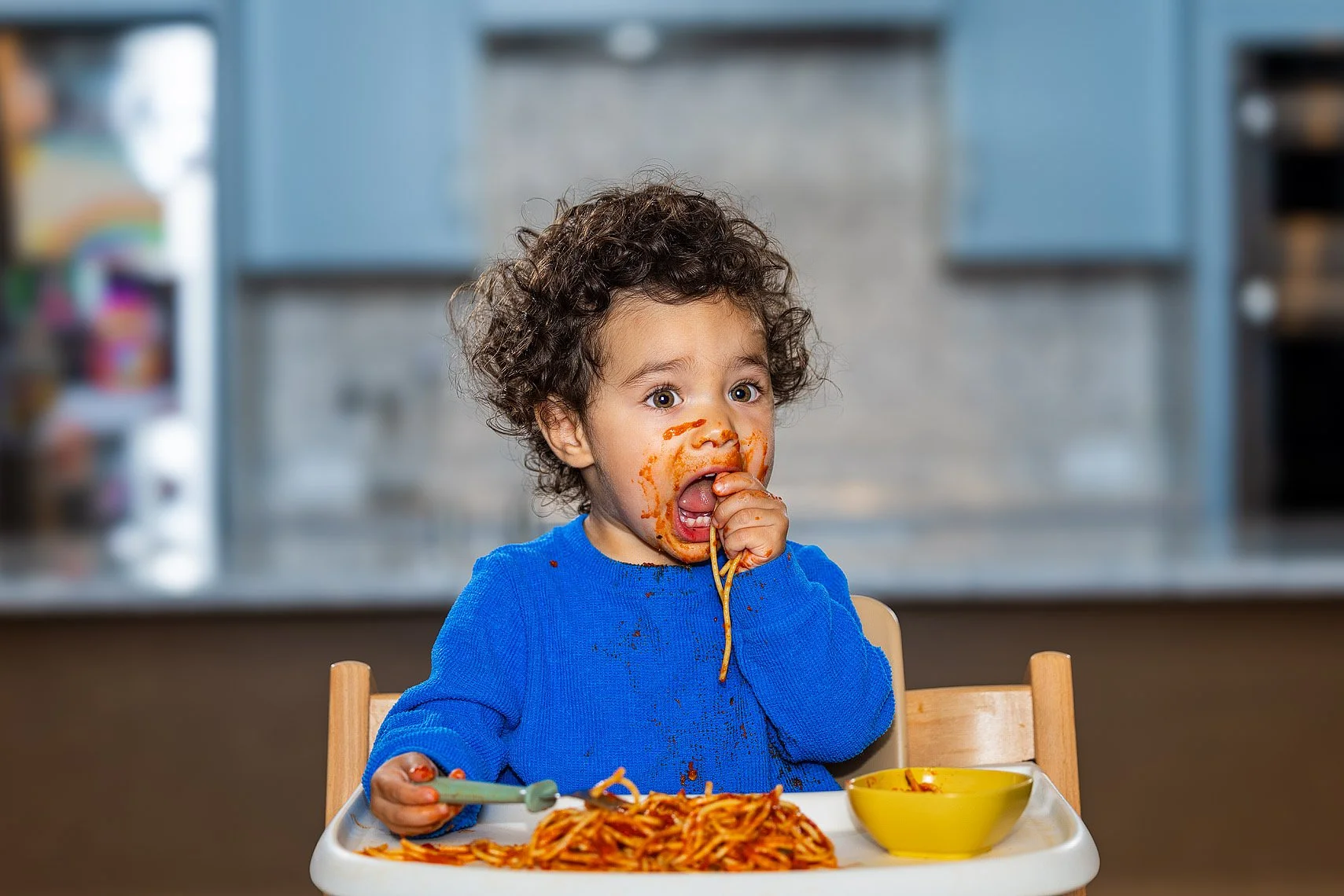 A young child with curly hair eating spaghetti, with sauce on face, sitting in a high chair at a table with a bowl of spaghetti.