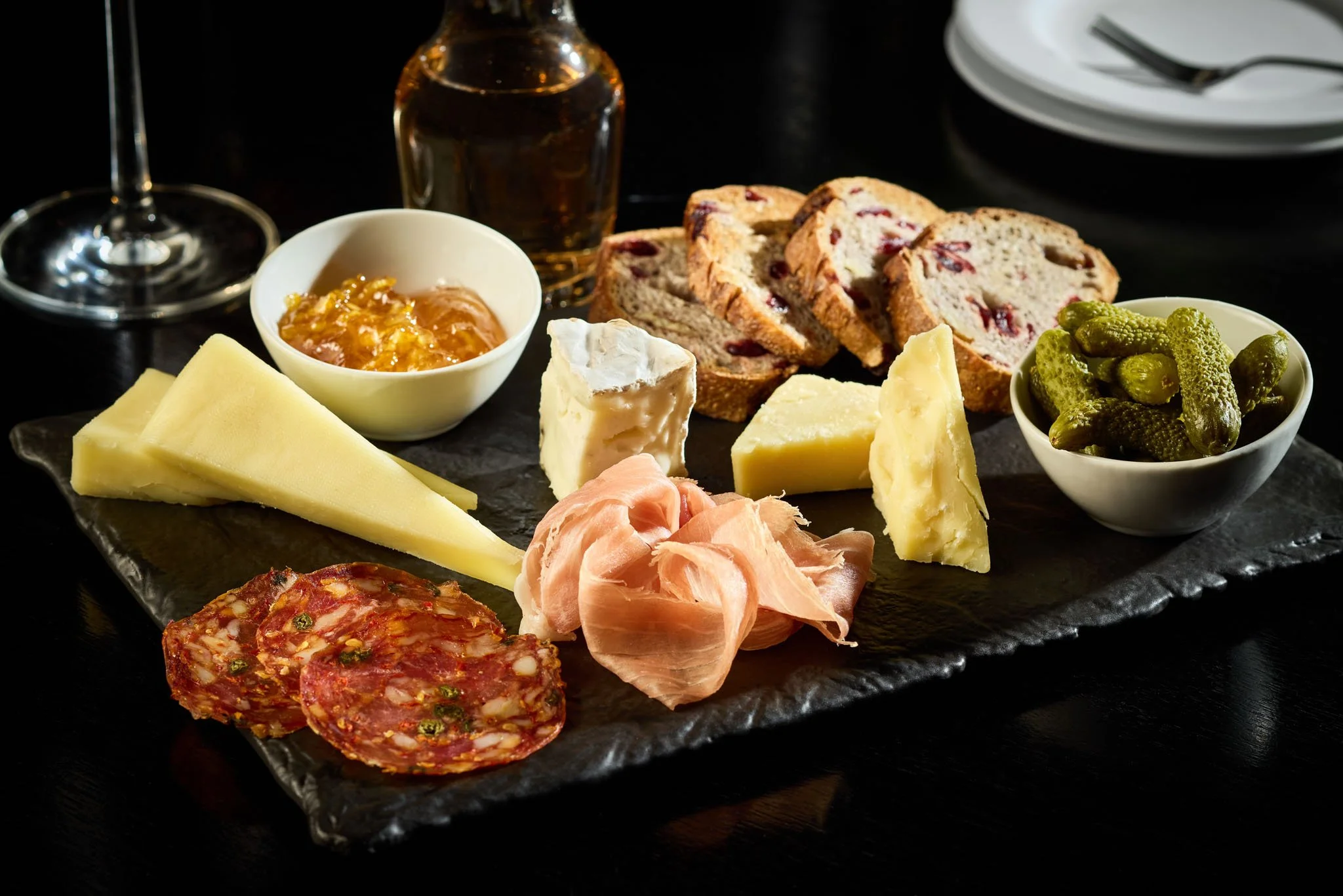 A cheese and charcuterie board with slices of cheese, cured meats, dried fruit, bread, and pickles, accompanied by a glass of red wine and a small bowl of jam on a black slate platter.