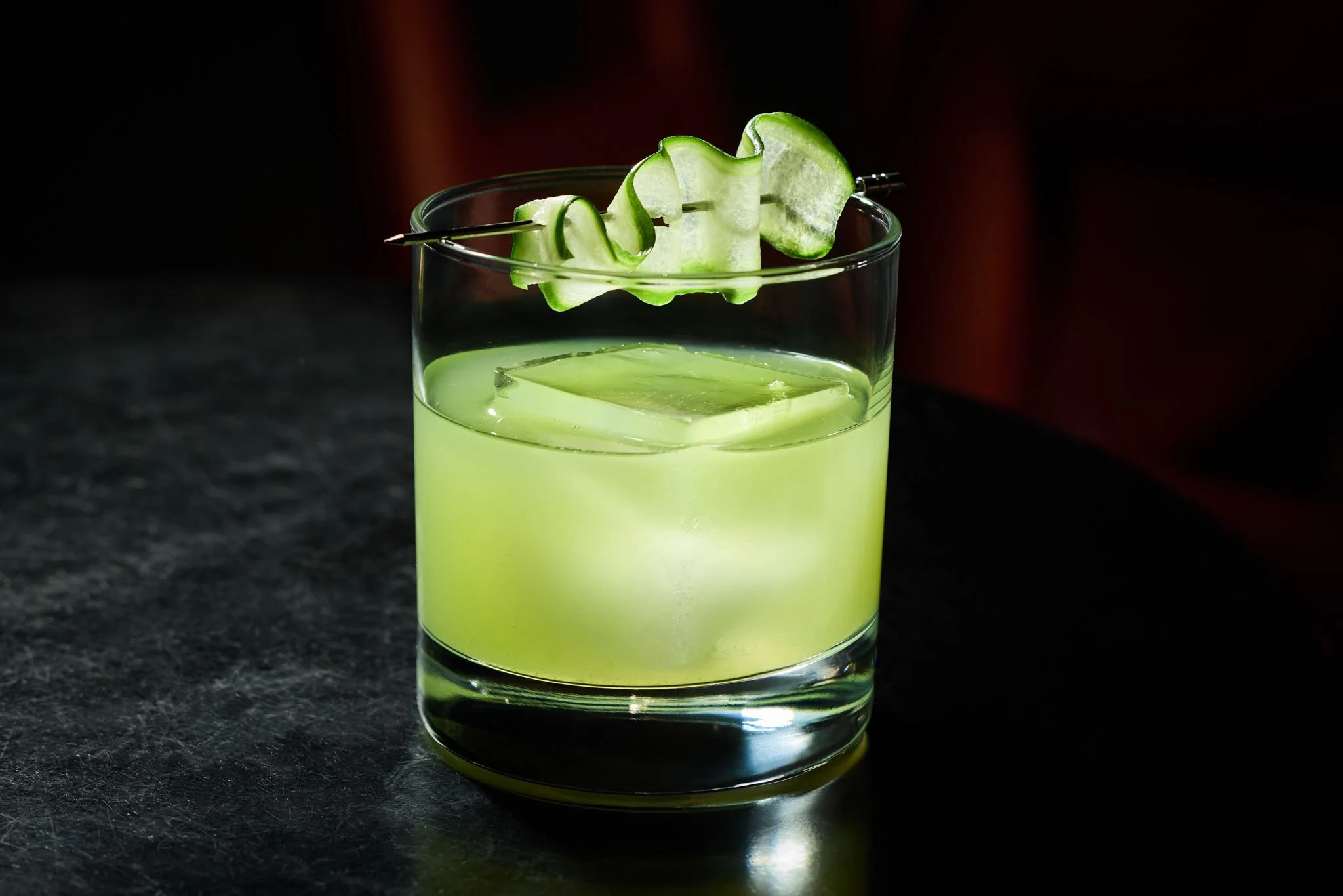 A glass of cucumber cocktail garnished with a cucumber peel twist on a cocktail pick, on a dark surface.