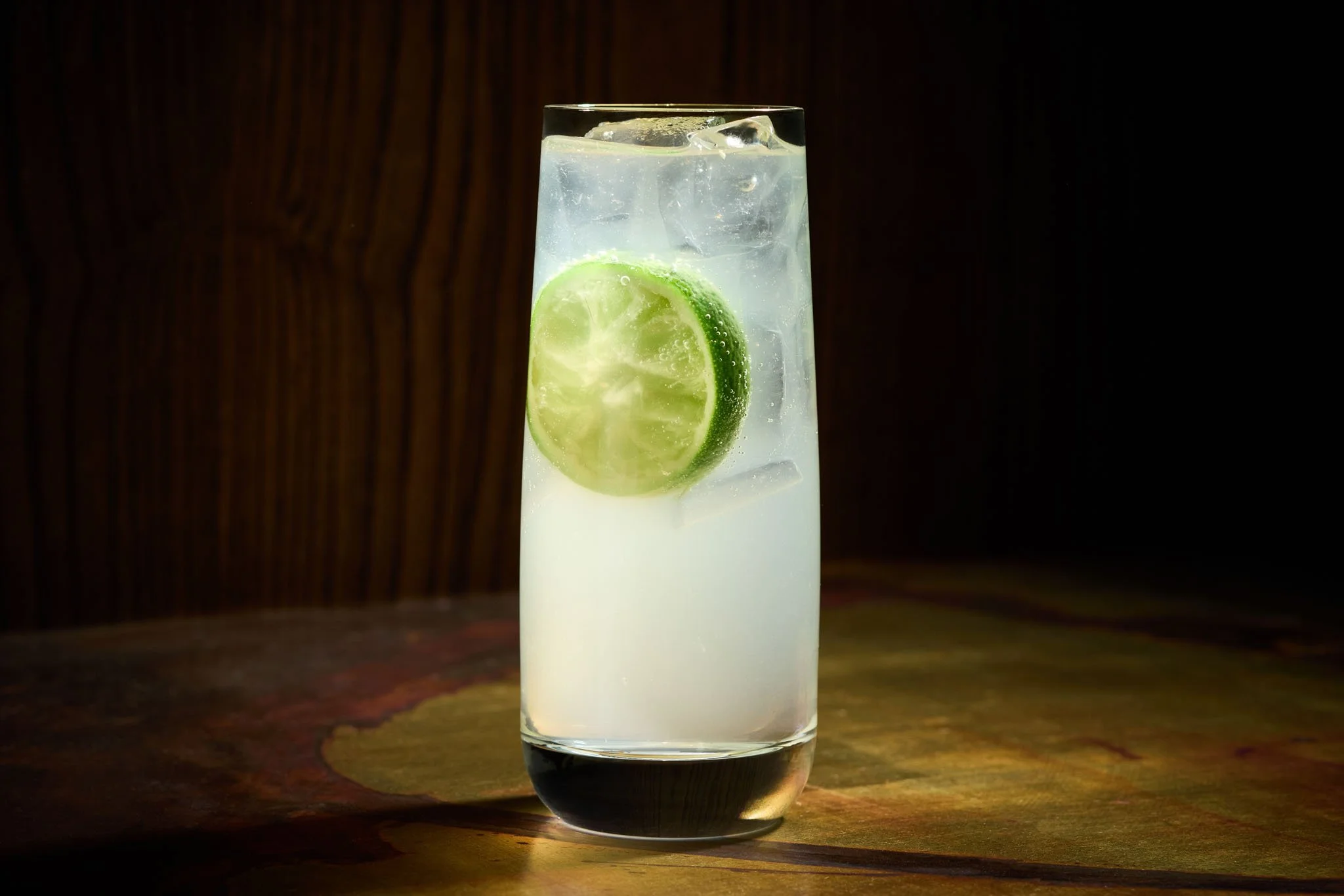 A tall glass of clear beverage with ice cubes and a slice of lime, set on a dark surface with a dark background.