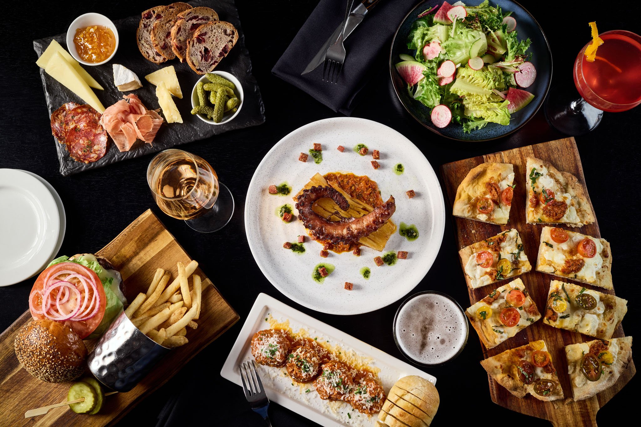 A spread of various dishes including cheese and charcuterie board, salad, pizza slices, a burger with fries, a wine glass with rosé, a bubbly beer, and a main plate with a piece of octopus with sauces on a dark table.