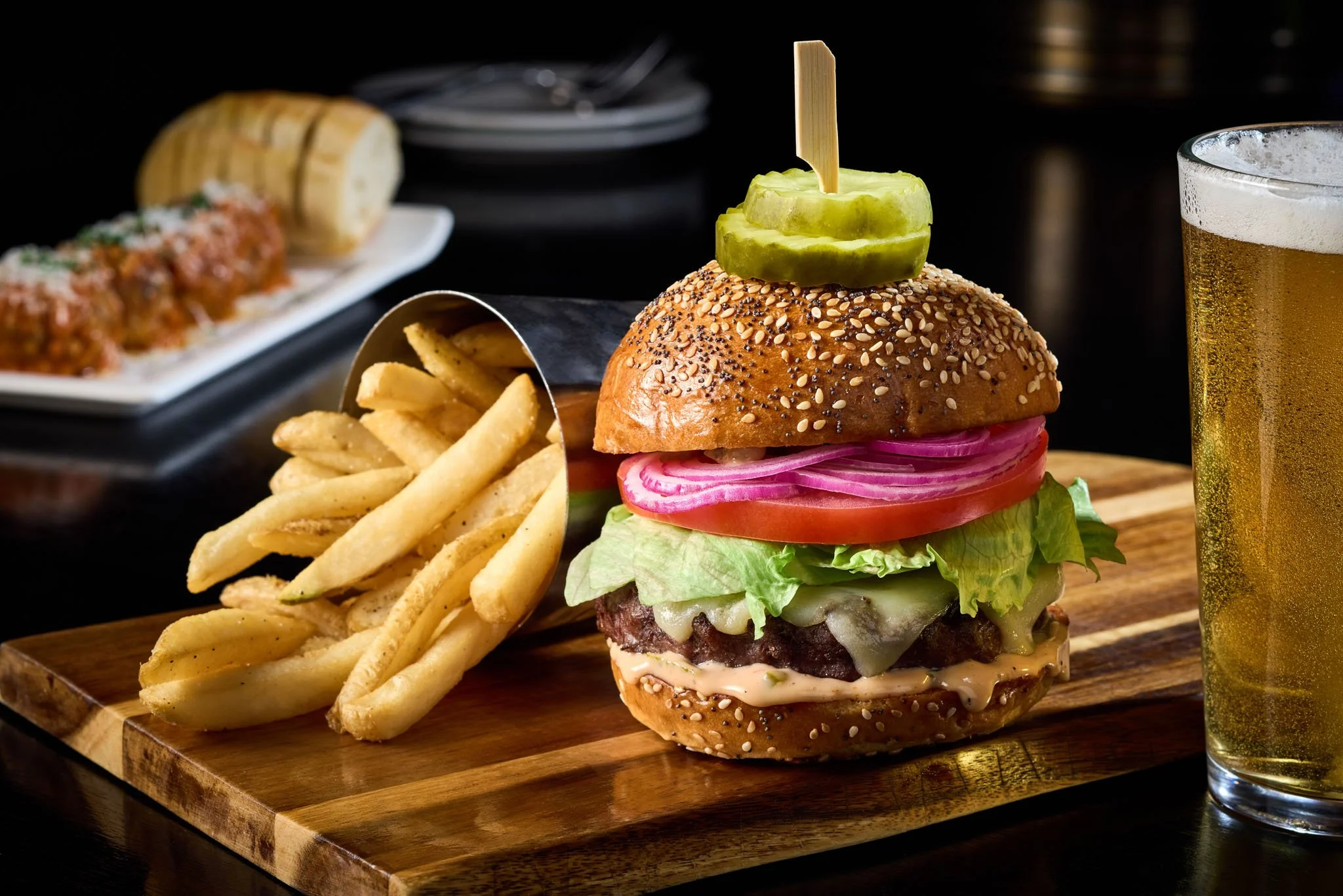 Juicy hamburger with lettuce, tomato, onions, cheese, and pickle on a sesame seed bun with fries and a glass of beer on a wooden board.