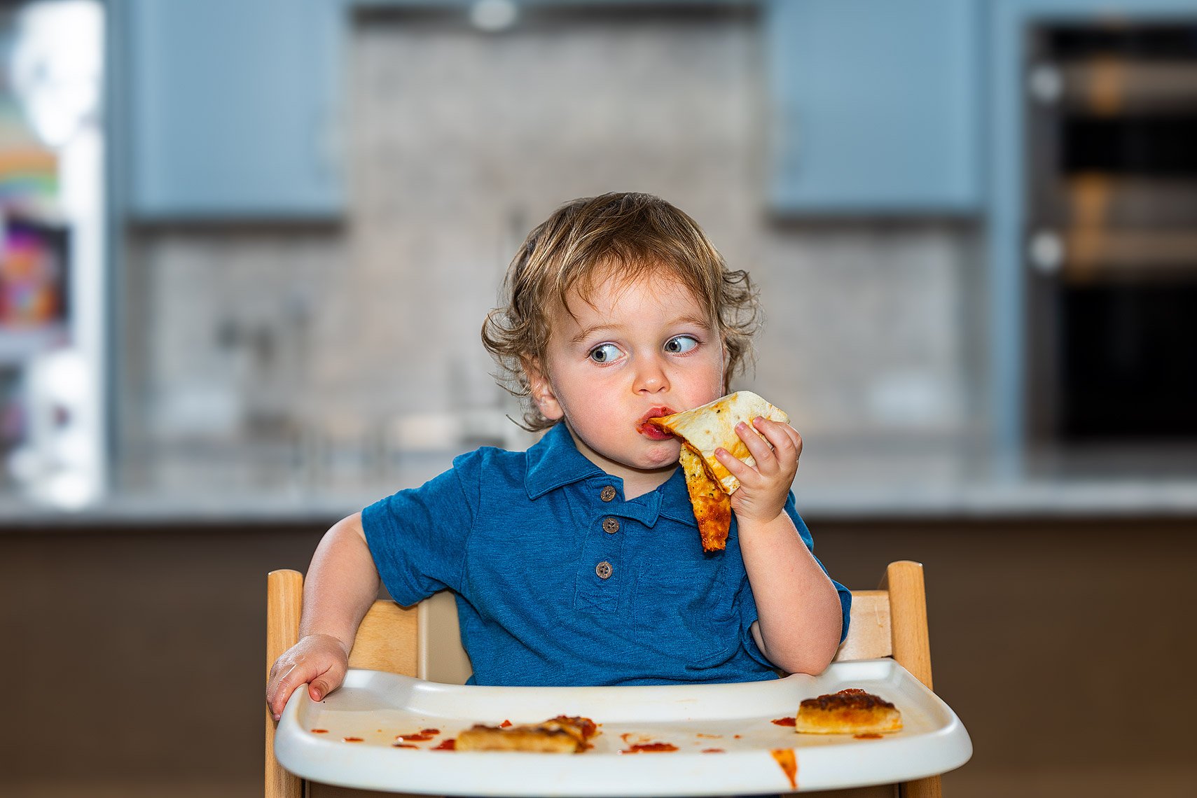 A young boy with curly hair and a blue shirt sitting in a high chair eating a slice of pizza with some remaining pizza on the tray.