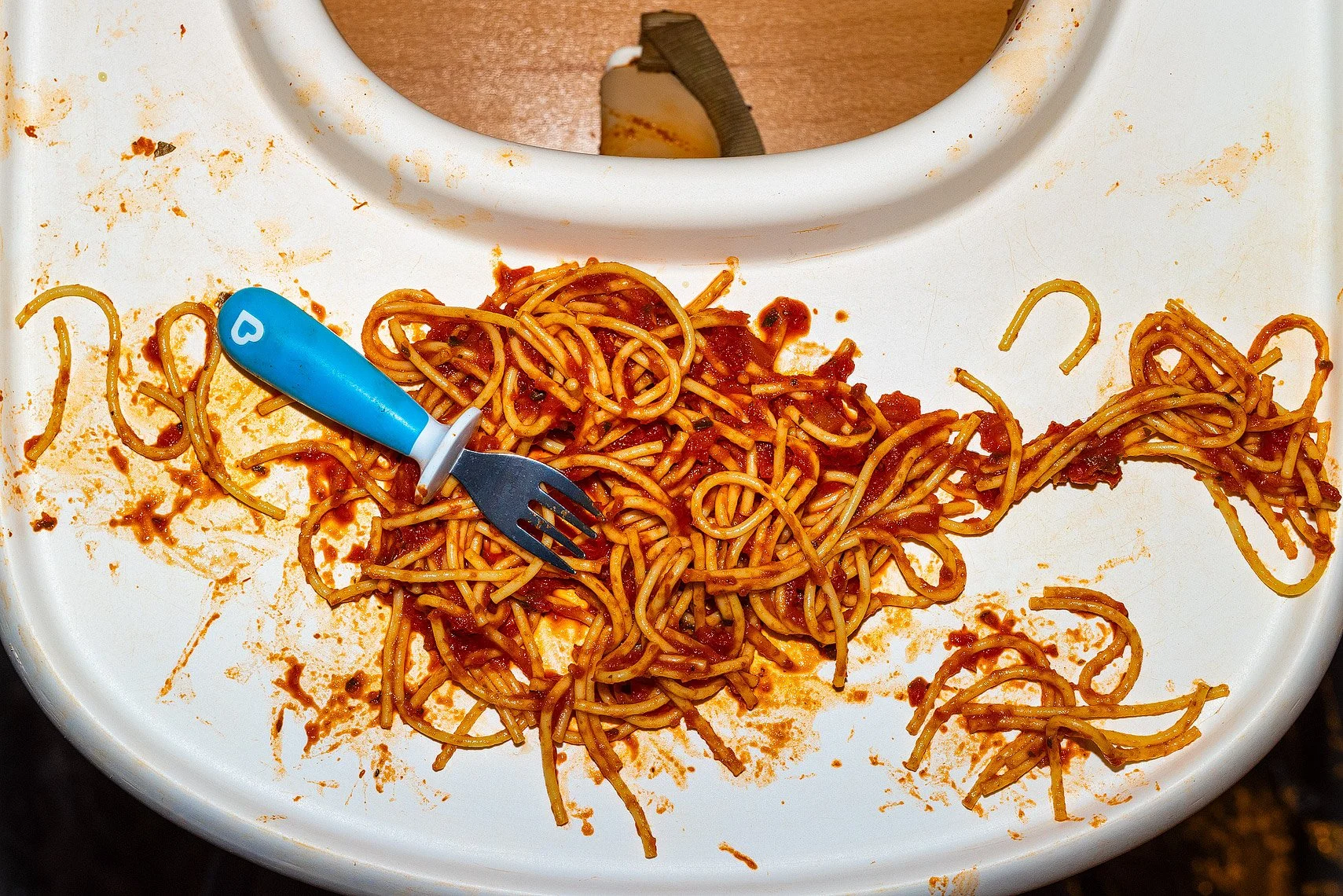 Spilled spaghetti with marinara sauce on a white high chair tray, a small blue child's fork resting on it, and a messy area with sauce splatters.