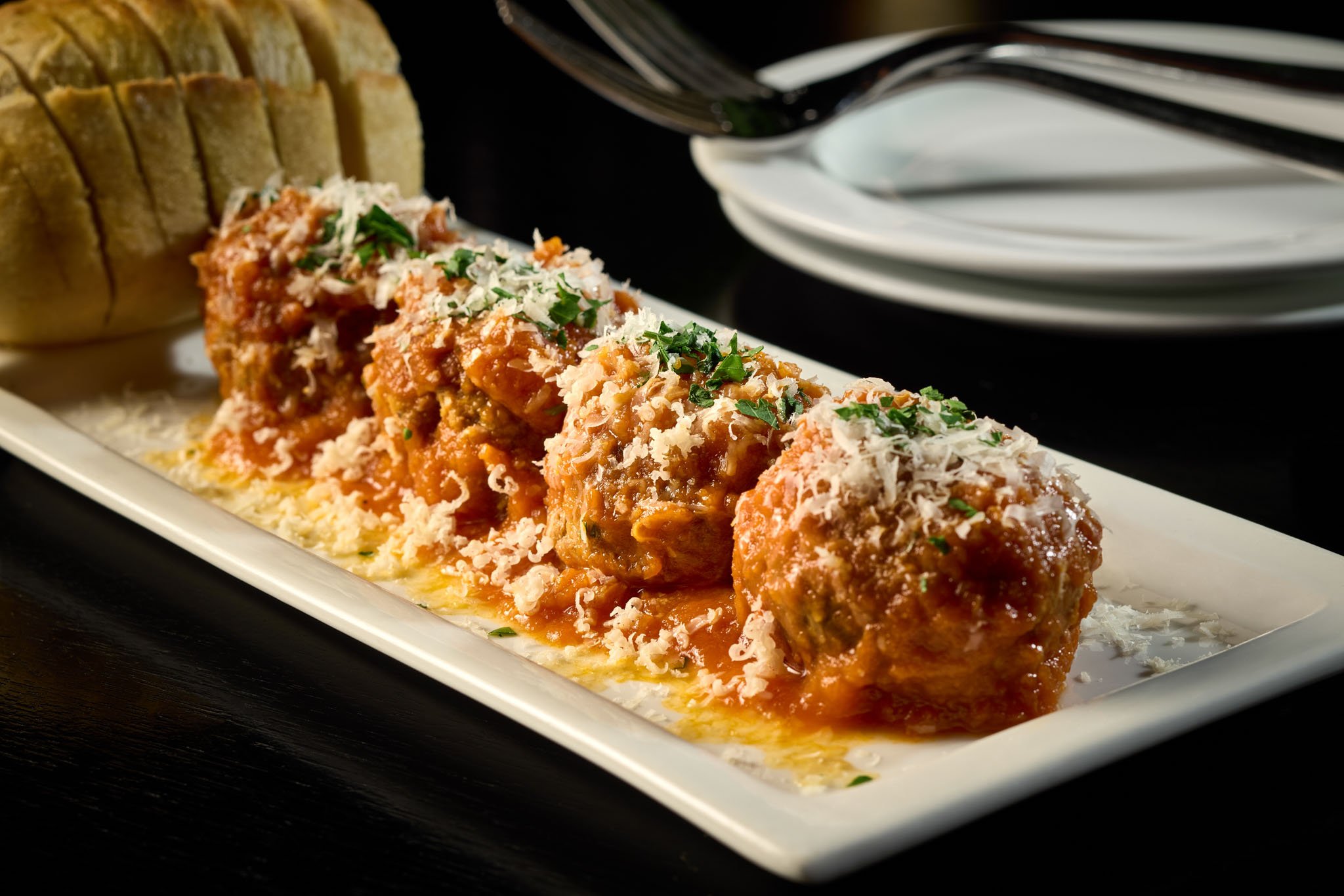 Plate of four large meatballs covered in tomato sauce, topped with shredded cheese and chopped herbs, with garlic bread slices in the background.