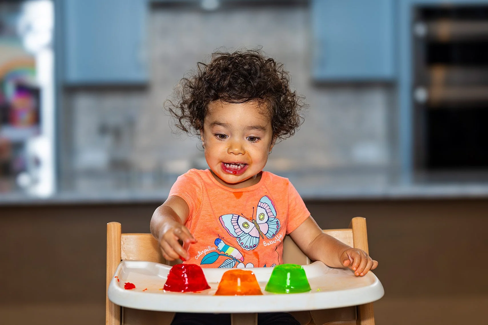 Child with curly hair in an orange shirt playing with colorful jelly desserts on a high chair tray.