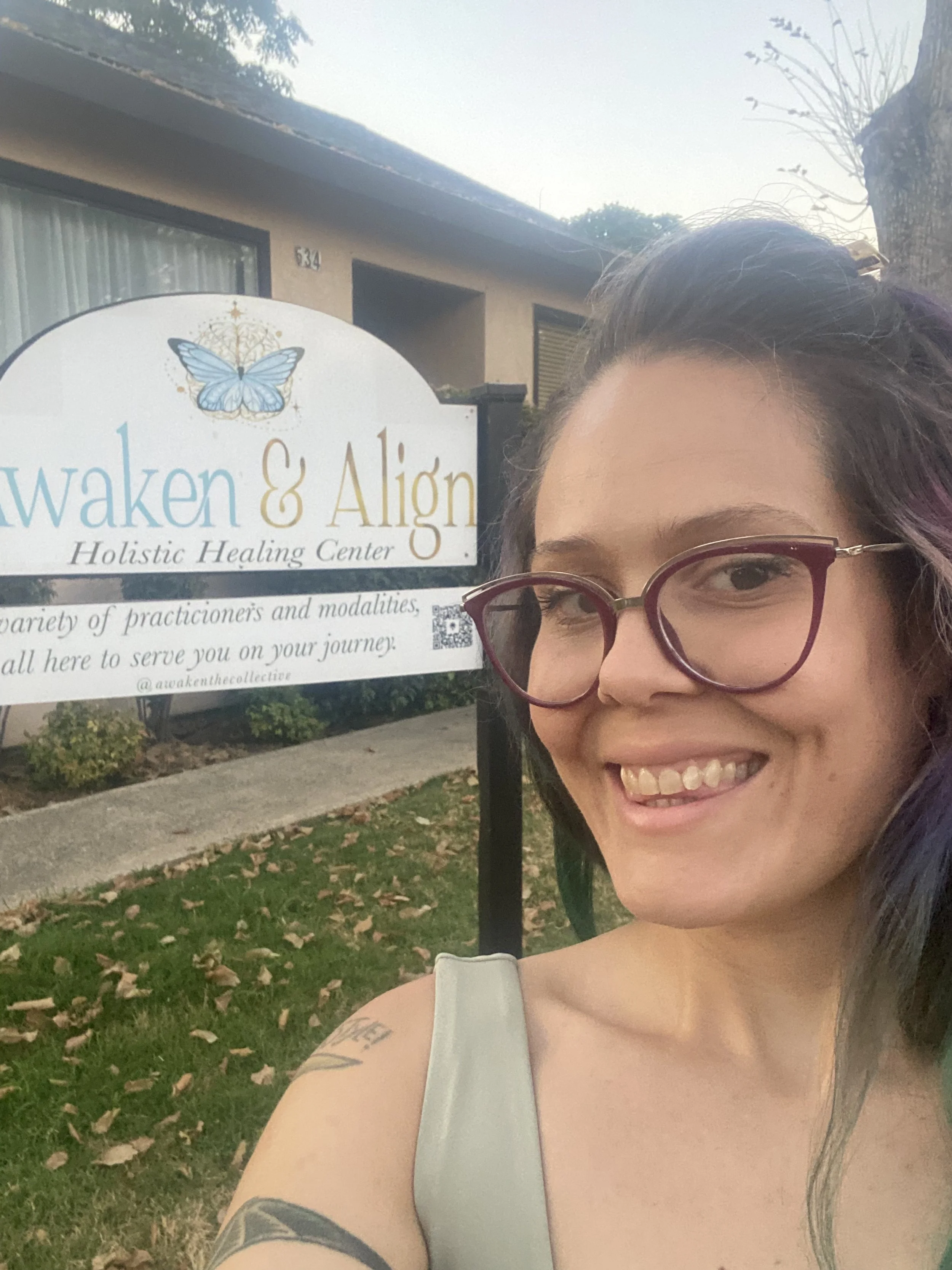 Smiling woman with glasses taking a selfie in front of a sign for 'Awaken & Align Holistic Healing Center' with a butterfly logo, in an outdoor setting with a building and trees in the background.