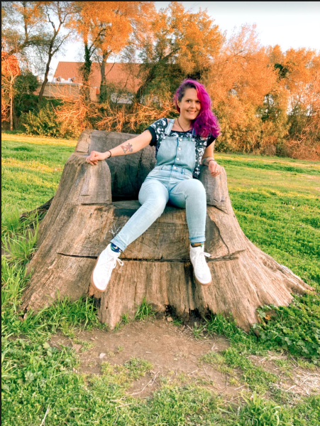 A young woman with long purple hair sitting on a large tree stump in a park during autumn. She is smiling and wearing a black t-shirt, denim overalls, and white sneakers.
