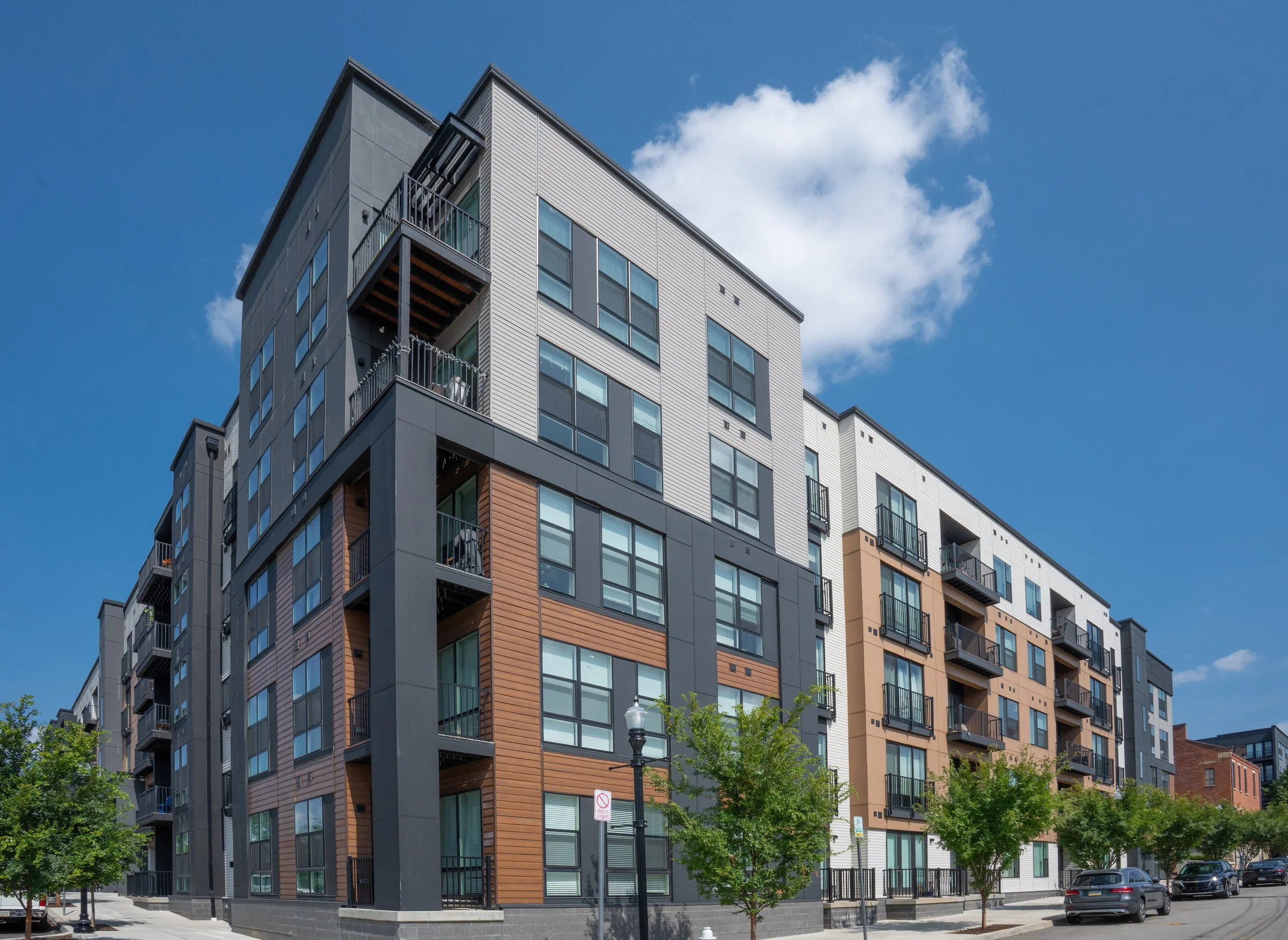 Modern apartment building with multiple floors and a contemporary design, featuring a mix of glass and wood paneling. The foreground shows a street with a pedestrian crossing sign and a streetlamp.