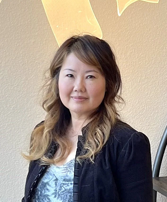 A woman with wavy, shoulder-length hair, wearing a gray shirt and black jacket, standing in front of a beige wall with a yellow decorative element.