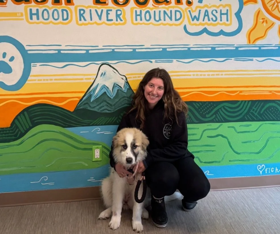A woman kneeling next to a fluffy puppy in front of a colorful mural of mountains, rivers, and text about a hound wash event.