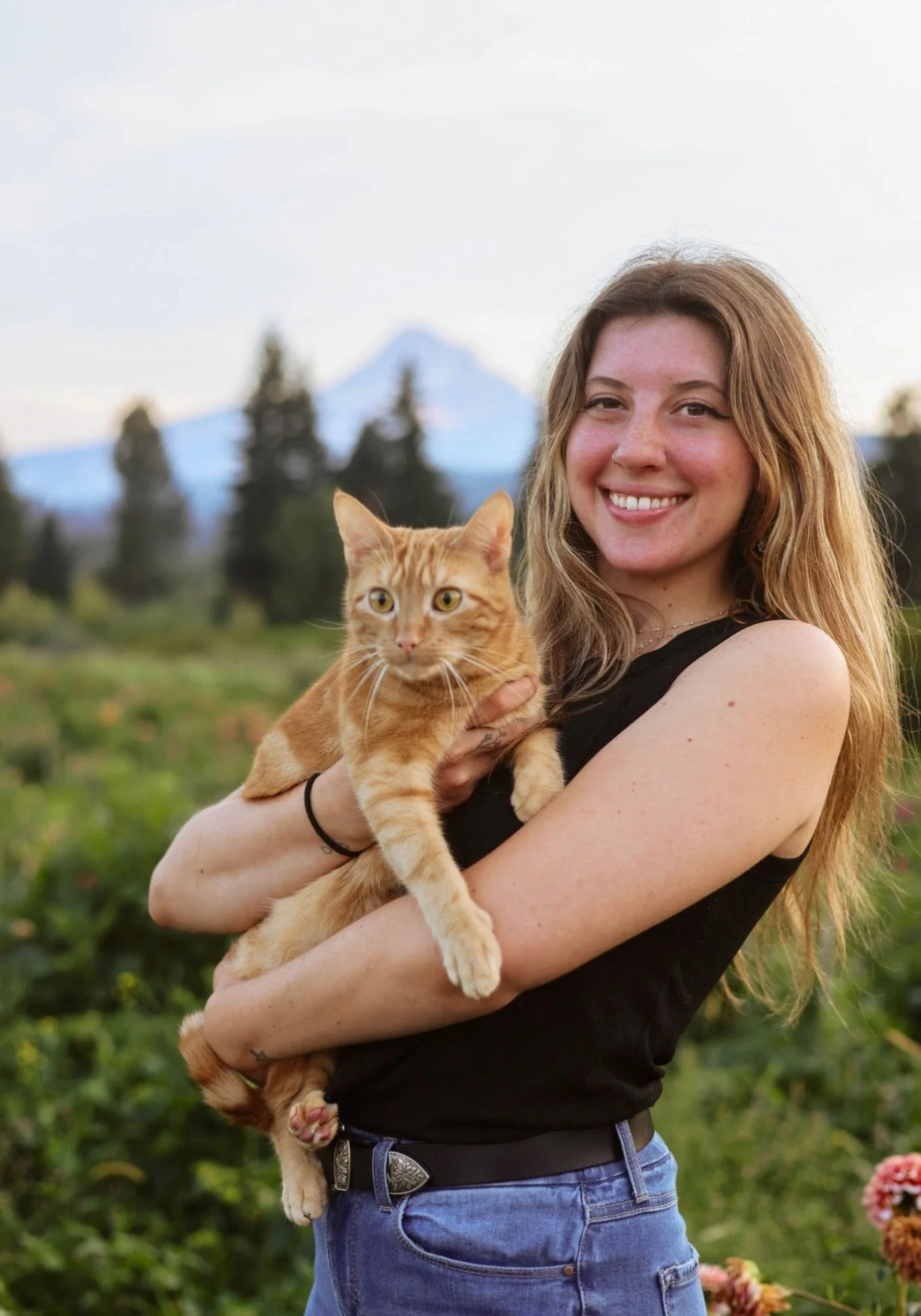 A woman with long, wavy reddish hair smiling and holding an orange tabby cat in a green outdoor setting with mountains and trees in the background.