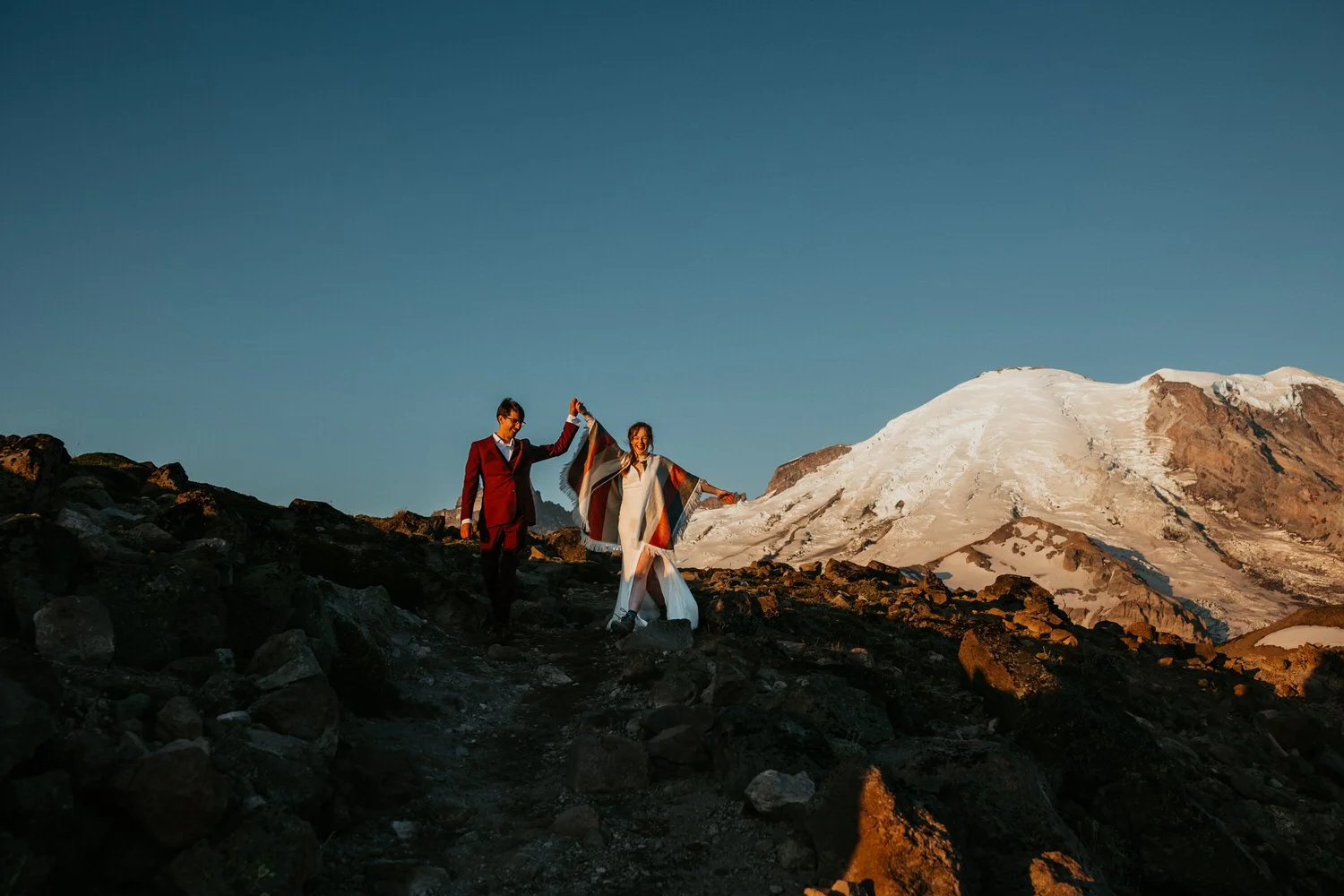 Couple hiking along a rocky trail at sunrise with Mount Rainier glowing in the background during a Washington elopement