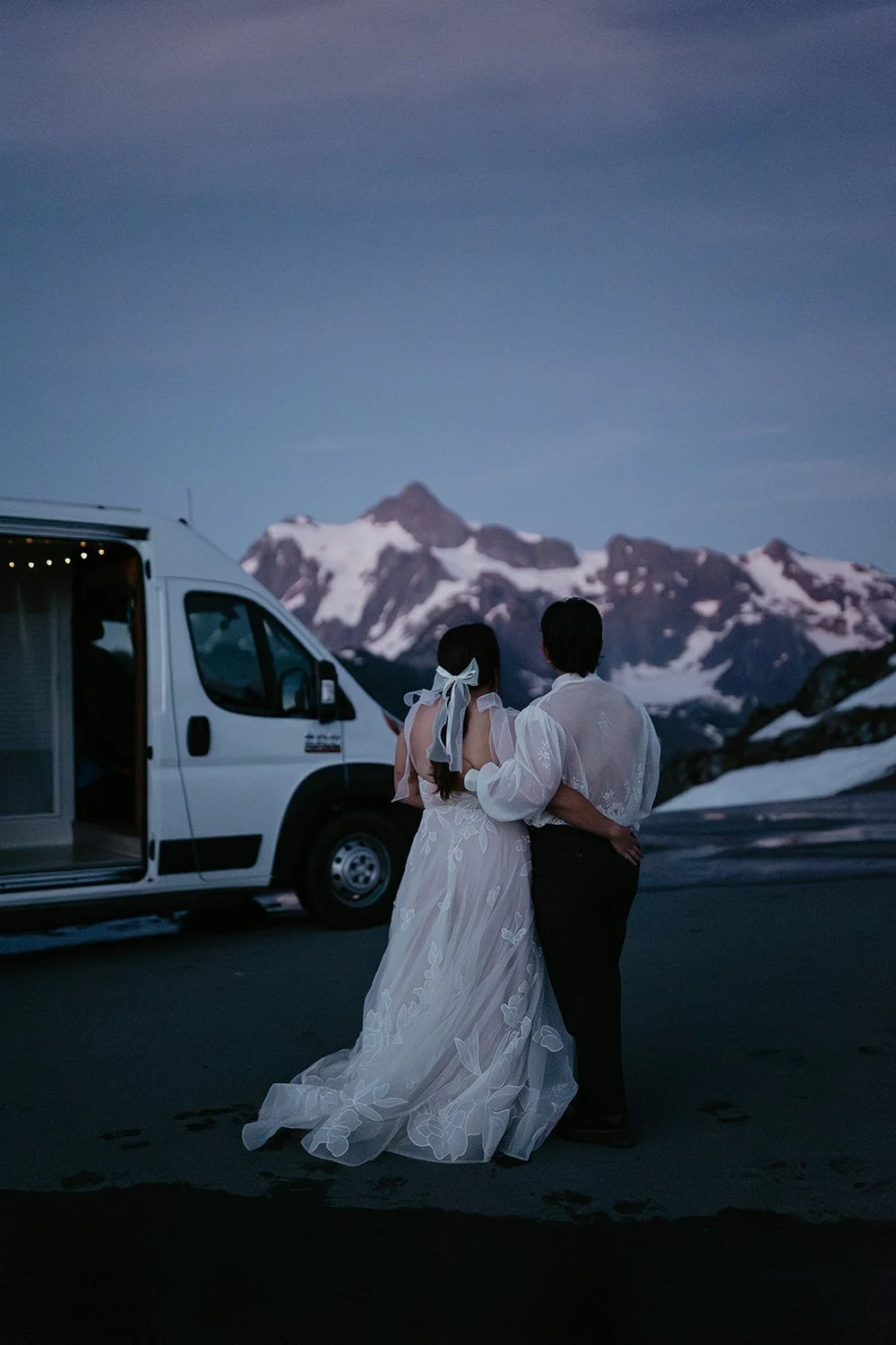 A couple stands arm in arm beside a camper van at dusk, looking out toward jagged, snow-covered mountain peaks under a deep blue sky.