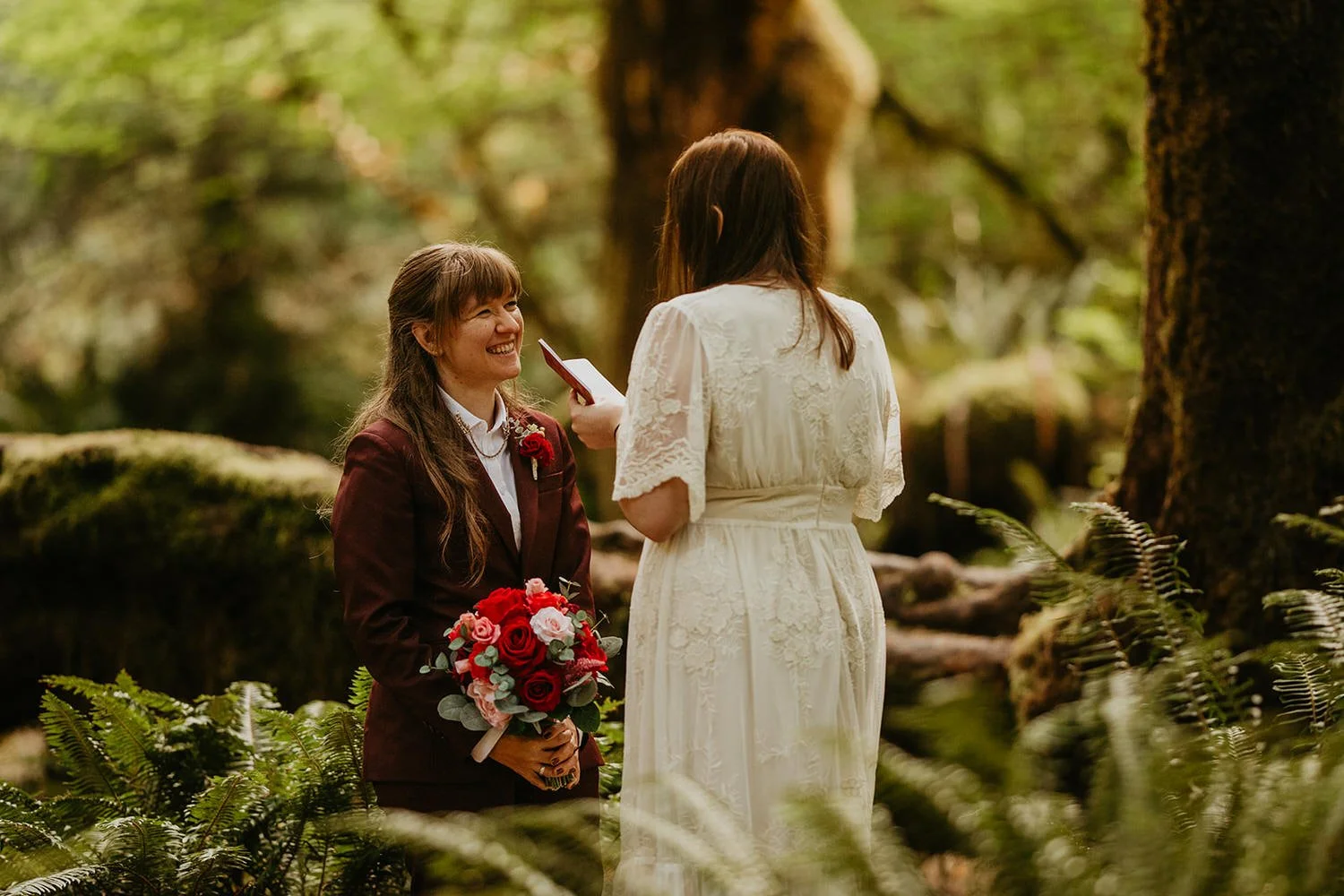 One bride reading vows to her partner during a forest elopement ceremony, surrounded by moss, ferns, and tall trees.