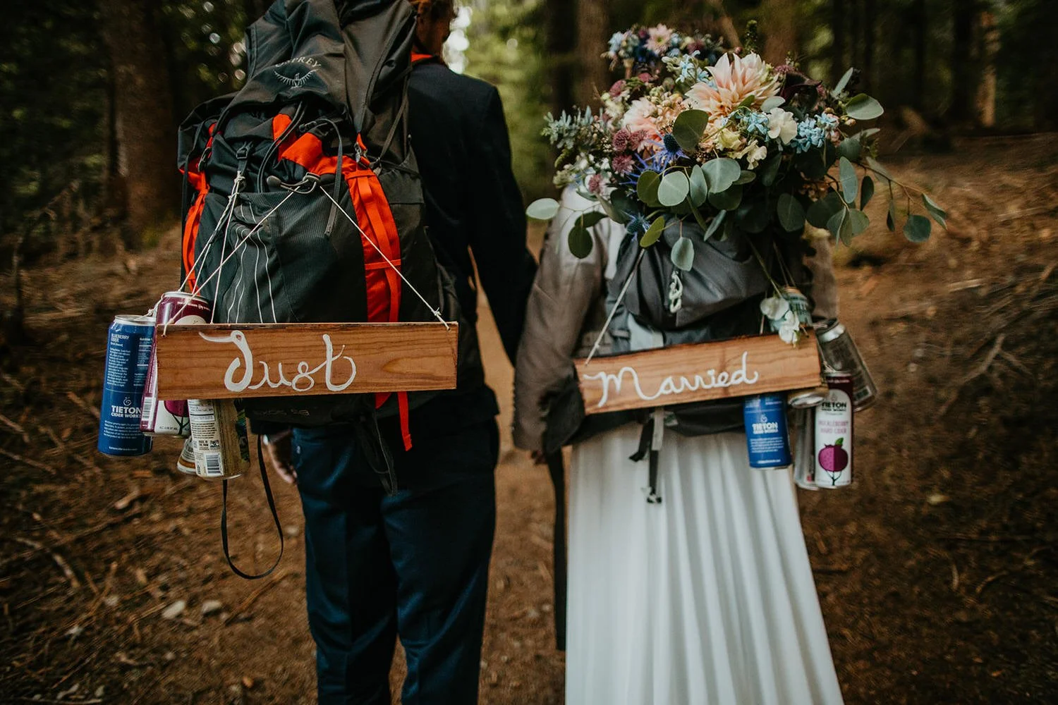 Backpacking newlyweds walking through a forest trail with Just and Married signs hanging from their packs.