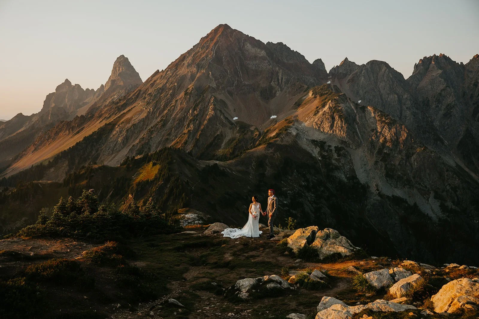 Couple standing on a rocky overlook in the North Cascades at sunset, jagged peaks glowing in warm light behind them during their Washington elopement.