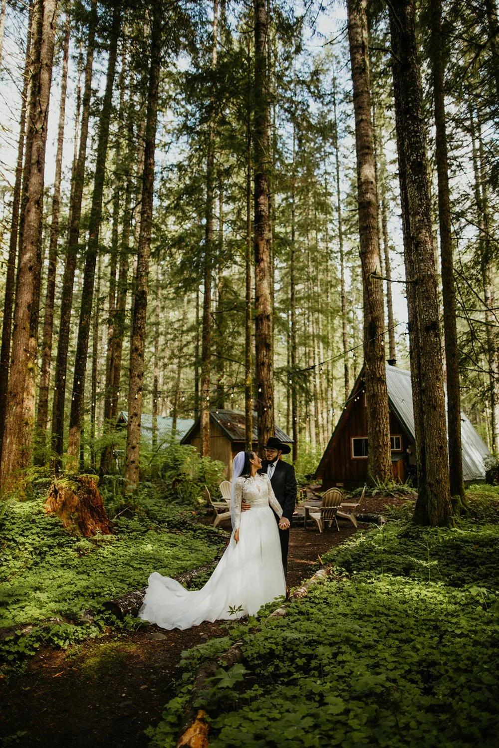 Couple in wedding attire sharing a kiss in a lush Pacific Northwest forest with a cabin visible through the trees