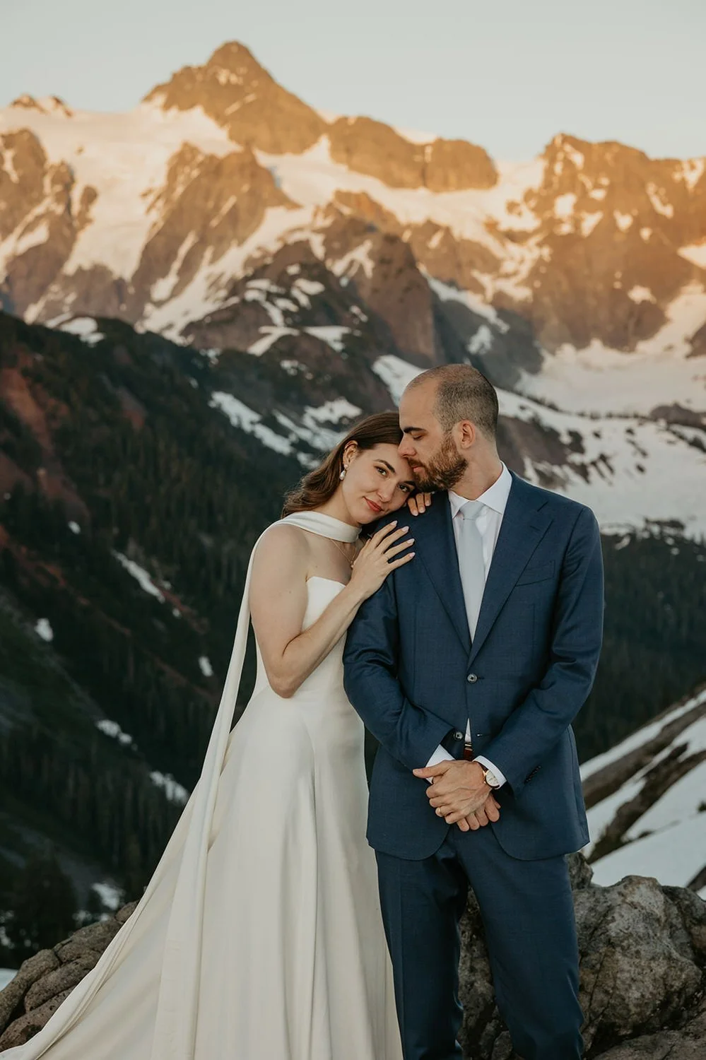 Couple embracing during a mountain elopement in Washington, standing on a rocky overlook with dramatic snow-covered peaks behind them.