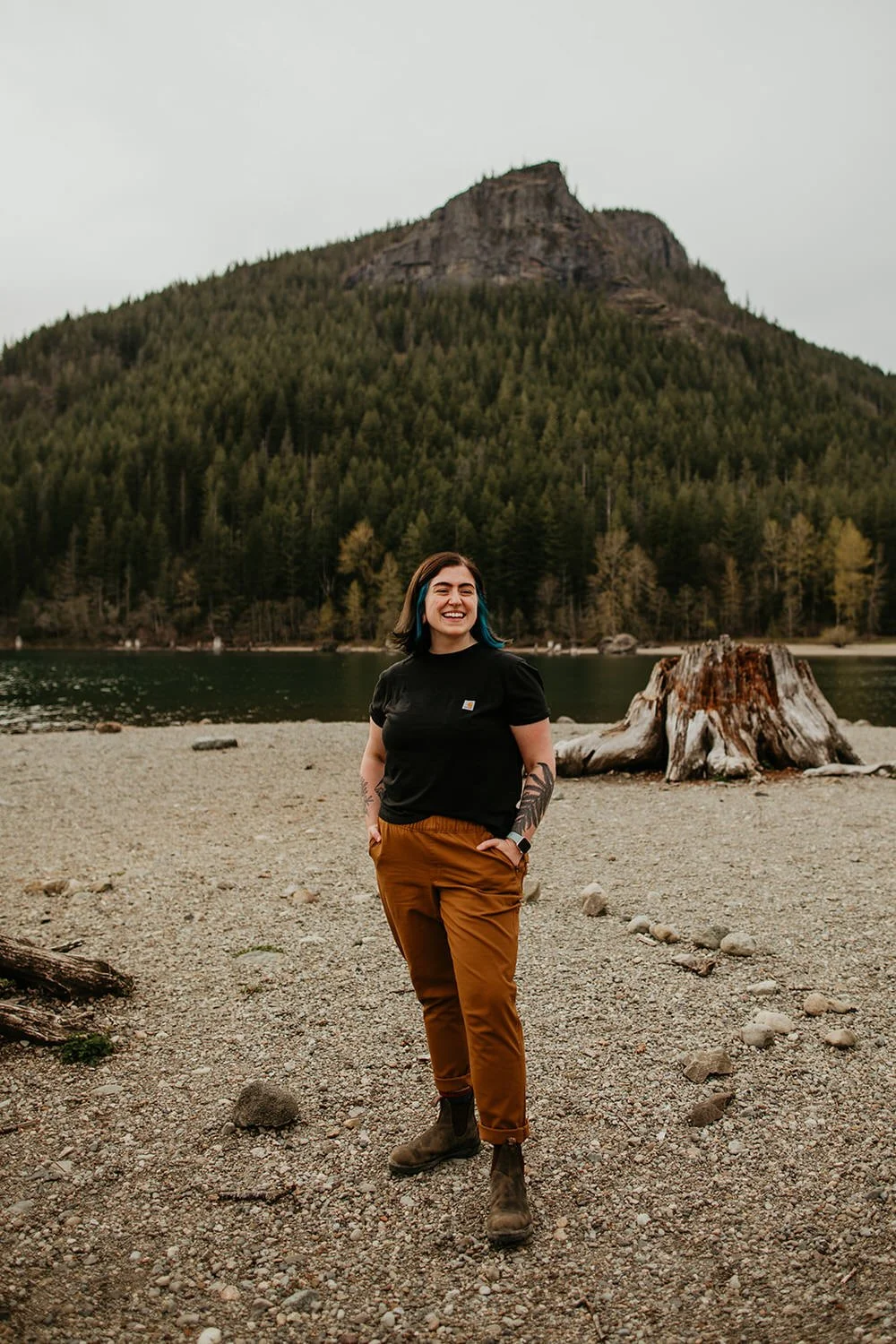 A woman standing on a rocky lakeshore, smiling, with a mountain and dense forest in the background.