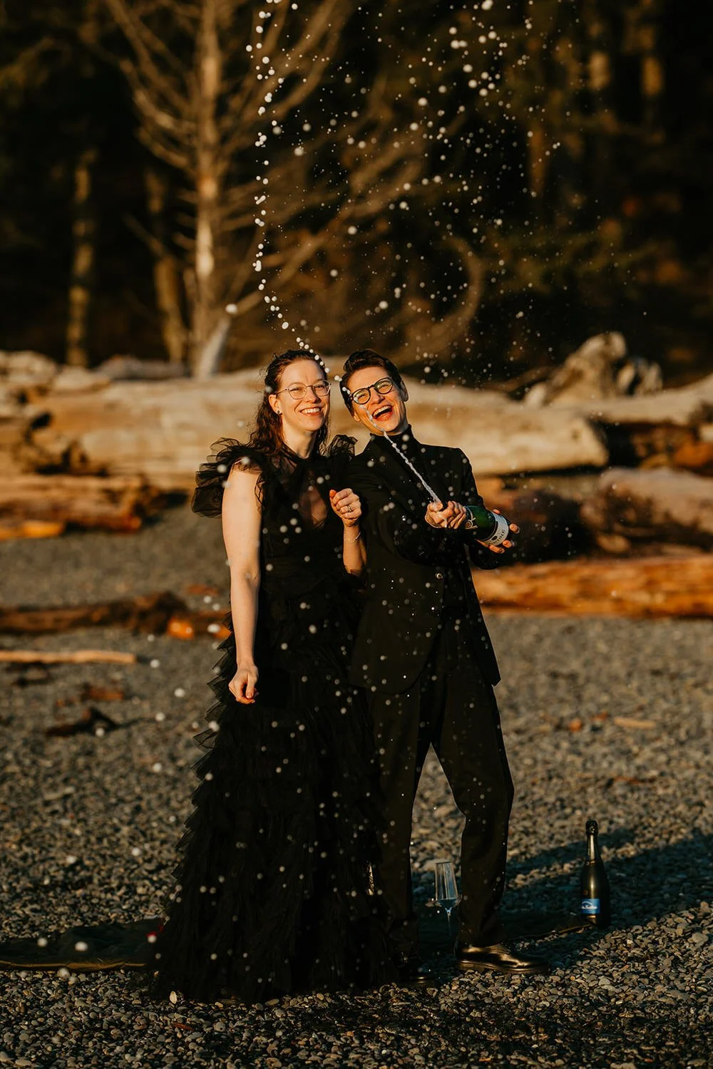 Two women in elegant black dresses celebrating on a rocky beach at night, opening a champagne bottle with champagne bubbles and spray in the air.