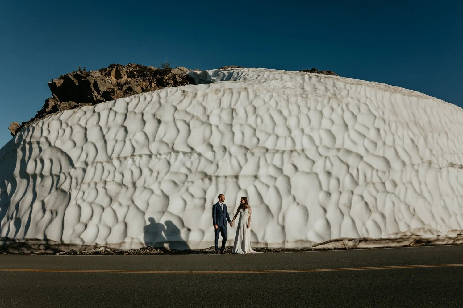 Couple holding hands beside a towering roadside snow wall along the Mount Baker Highway in early summer