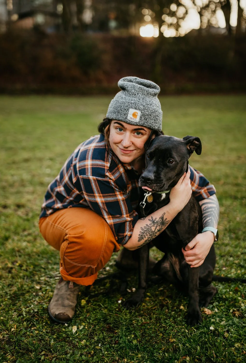 A woman crouches on grass during sunset, hugging a black dog, both looking content. She is wearing a gray Carhartt beanie, a plaid shirt, orange pants, and brown boots, with tattoos on her arm.
