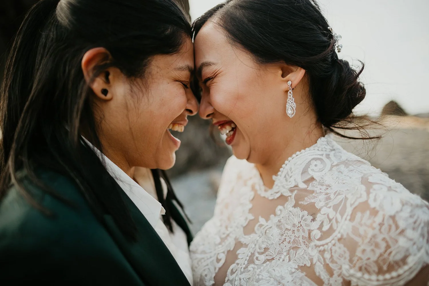 Two brides laughing with their foreheads touching during an intimate outdoor elopement, one wearing a lace wedding dress and the other in a dark suit.
