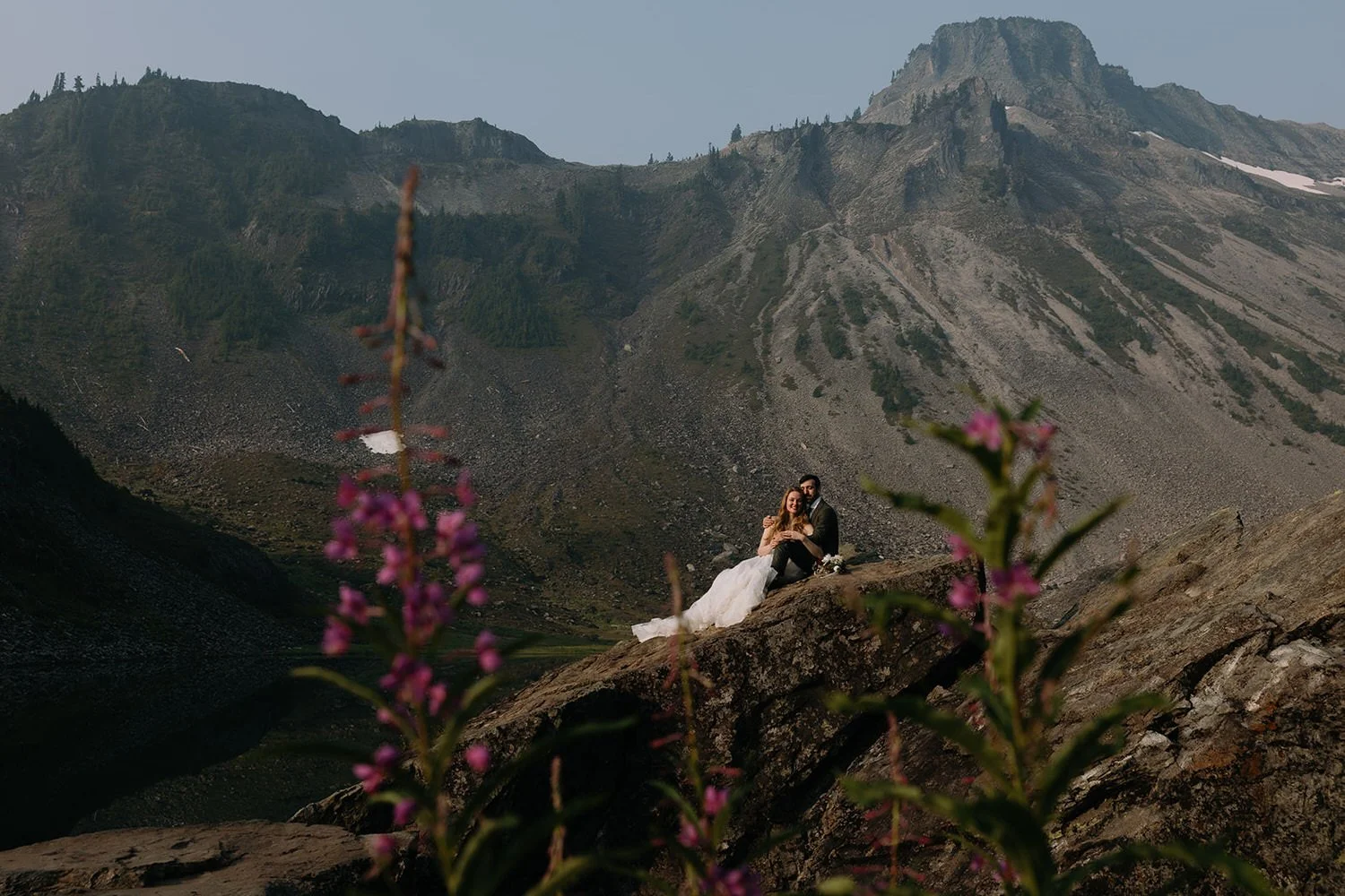 Couple sitting on rocky outcrop overlooking volcanic landscape with pink wildflowers in sharp foreground with artist point in the background